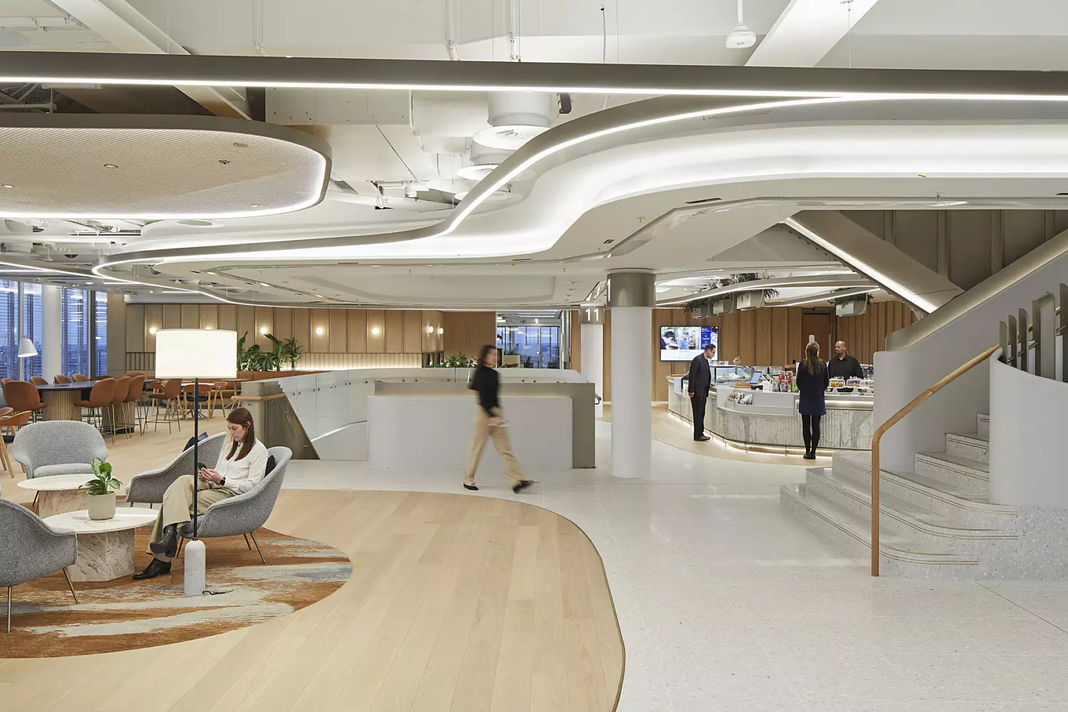 Office interior with a spacious, open-plan layout featuring multiple seating areas furnished with contemporary chairs and tables. The ceiling is fitted with distinctive curved lighting fixtures, and a staircase on the right leads to an upper level. The flooring combines light wood and white tiles, while large windows on the left bring in ample natural light. Several people are visible, either walking through the space or seated in conversation.