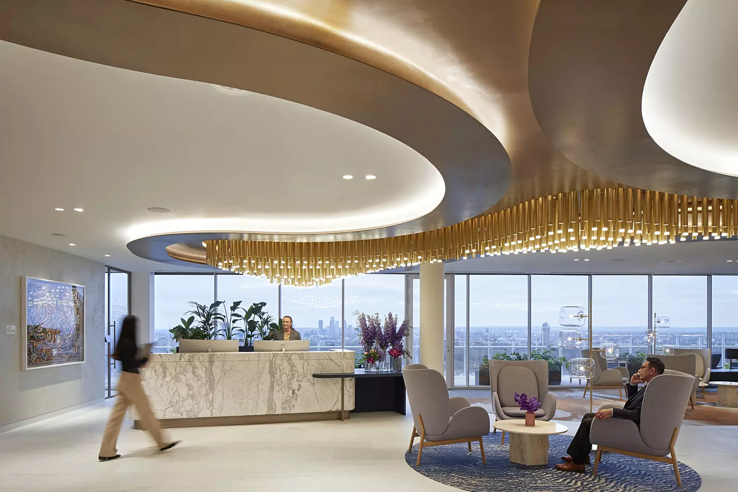 Modern office lobby with a refined and elegant design, featuring a reception desk on the left and a large curved ceiling installation adorned with hanging golden elements. The space includes a seating area with two chairs and a small table topped with flowers, creating a welcoming atmosphere. Large windows at the back offer views of an urban skyline, while the overall design blends luxury with contemporary minimalism.