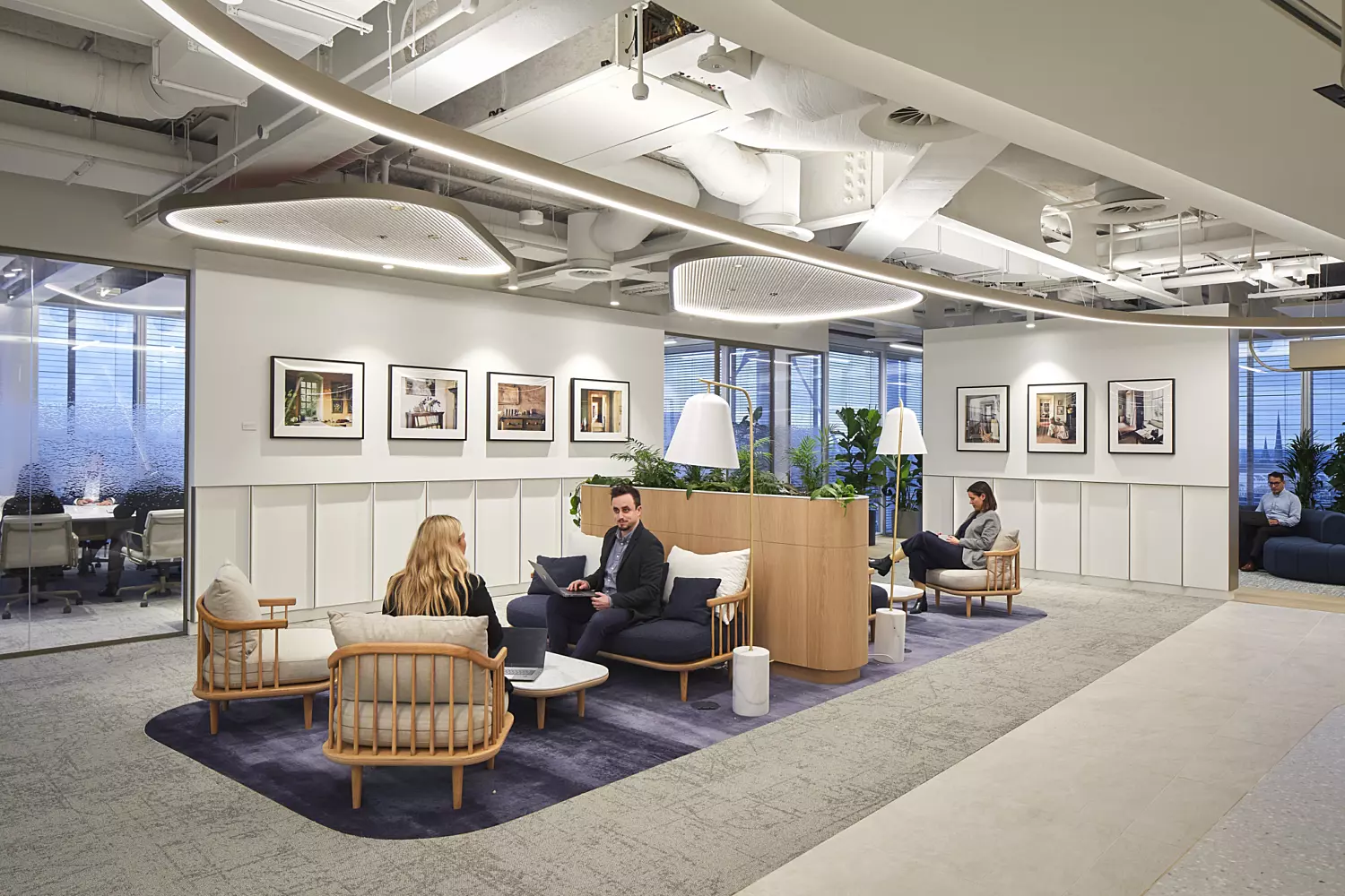 Office lounge area with a clean, contemporary design, featuring three people seated on chairs and sofas around a small table. The space is illuminated by ceiling lights and natural light from large windows in the background. White walls are decorated with framed pictures, and potted plants add a touch of greenery, creating a relaxed and welcoming environment.