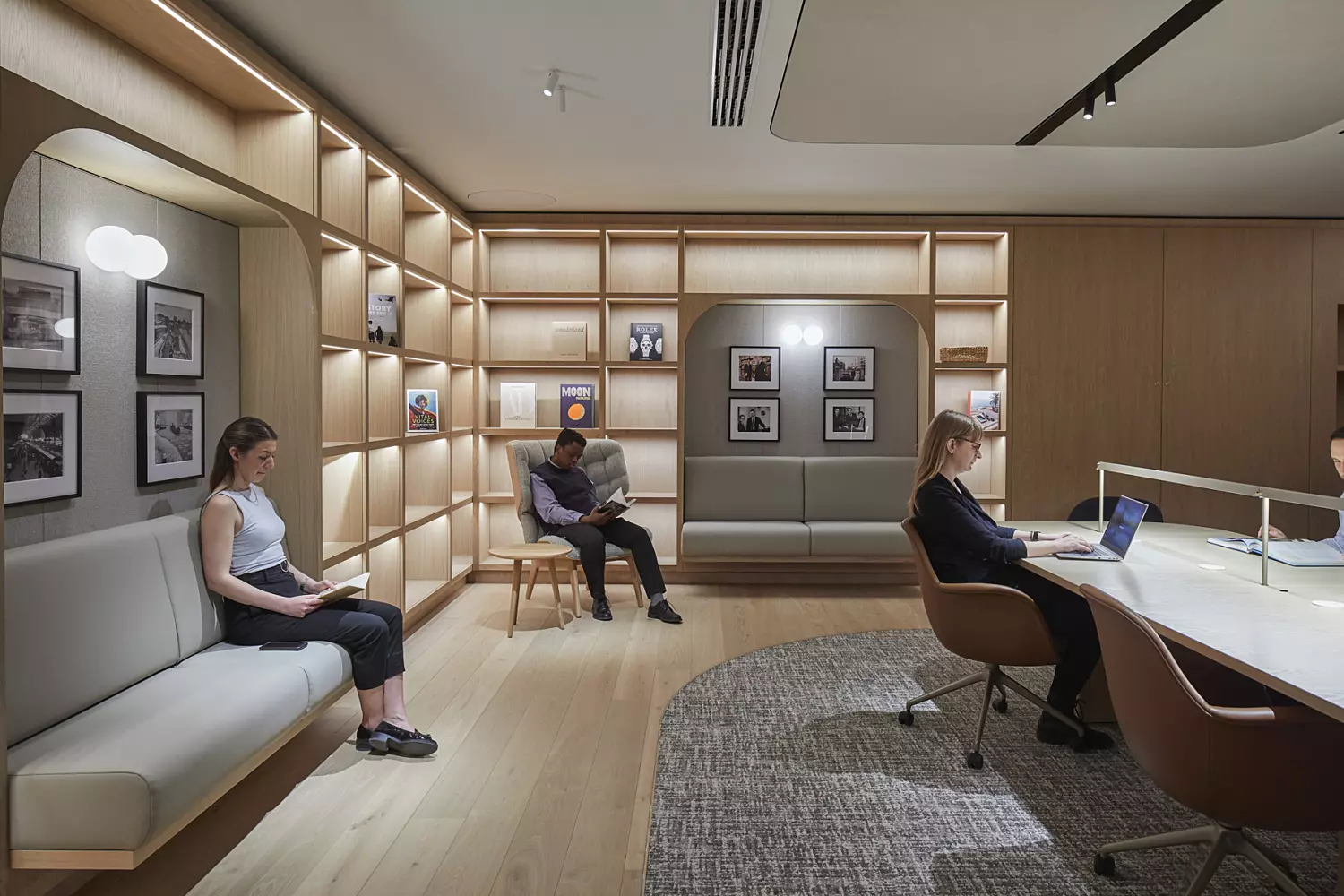 Office space with a warm, inviting atmosphere, featuring built-in wall shelves filled with books and framed pictures. The room includes a mix of seating options, such as cushioned benches along the wall and chairs arranged around a large central table. Soft, ambient lighting enhances the comfortable and collaborative working environment.