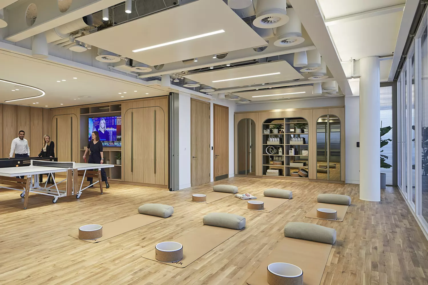 Spacious casual office area with wooden flooring and a mix of open and enclosed zones. On the left, a group of people are gathered around a ping pong table, while the central and right sections feature floor cushions and mats, suggesting a space for relaxation or meditation. The back wall is lined with shelves holding books and decorative items. The ceiling features an intricate panelled design with integrated lighting, and large windows on the right bring in generous natural light