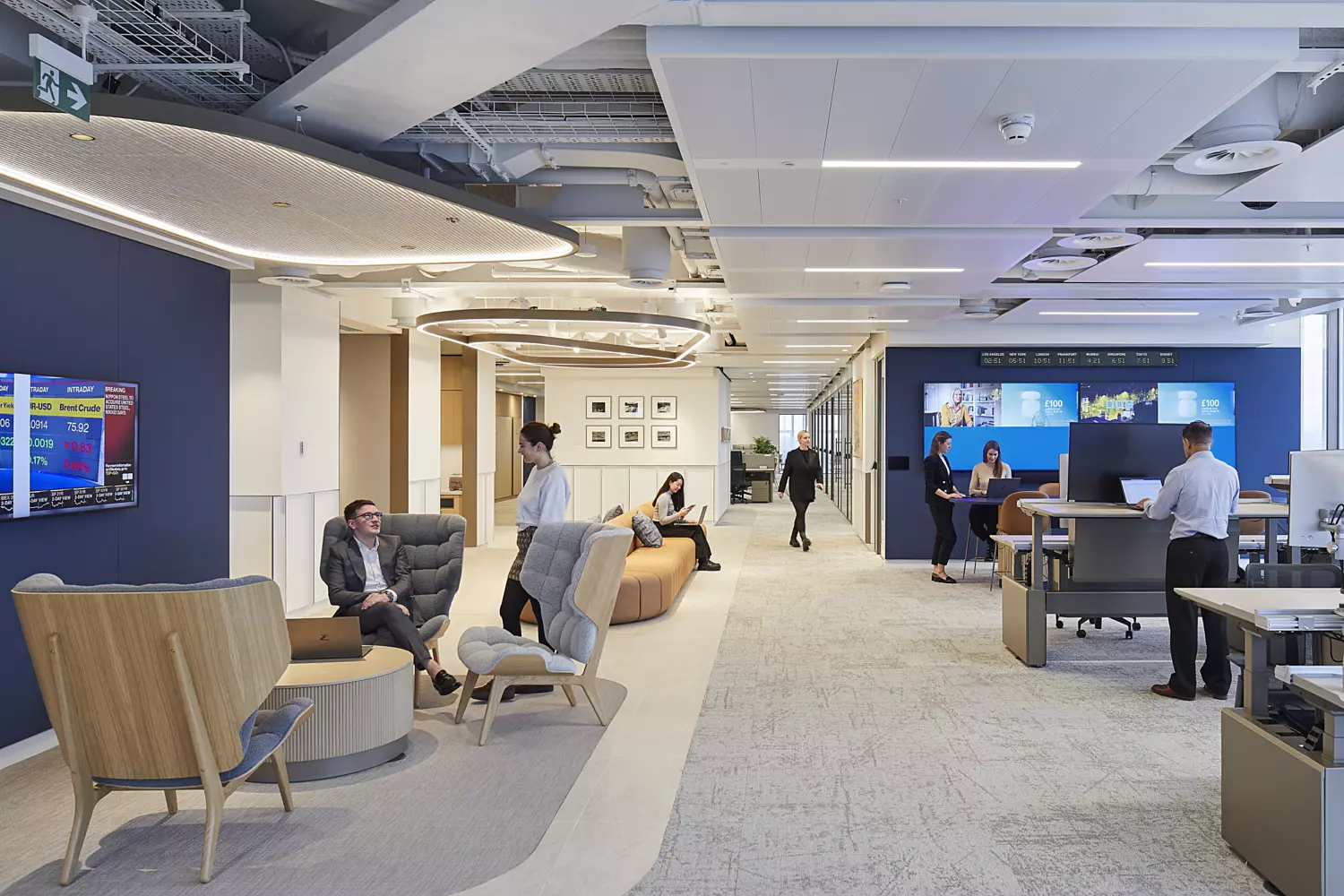 Modern office space with a contemporary layout featuring blue walls, exposed ceilings, and well-distributed lighting. The environment includes a mix of seating options such as chairs, couches, and desks equipped with computers. Multiple wall-mounted screens display various content, contributing to a dynamic and tech-forward atmosphere designed to support both collaborative and individual work.