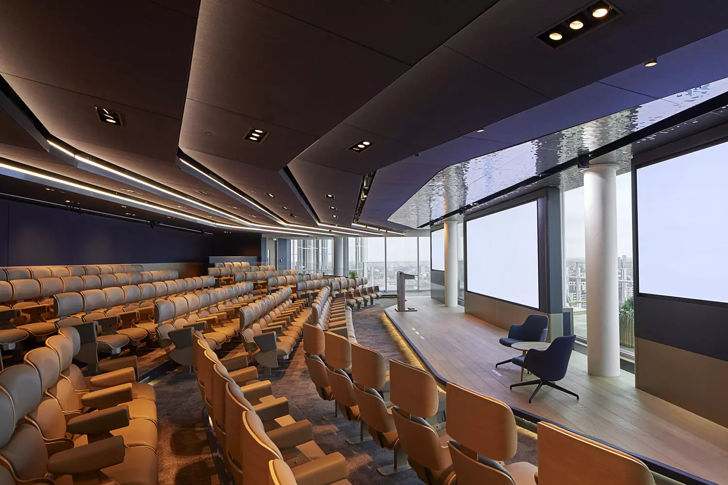 Modern auditorium-style conference space with rows of empty seats arranged to face a stage area. The stage includes two chairs and a podium, set against a backdrop of large windows that allow natural light to fill the space. The ceiling features recessed lighting, and the overall design is sleek and contemporary, combining functionality with a clean, professional aesthetic.