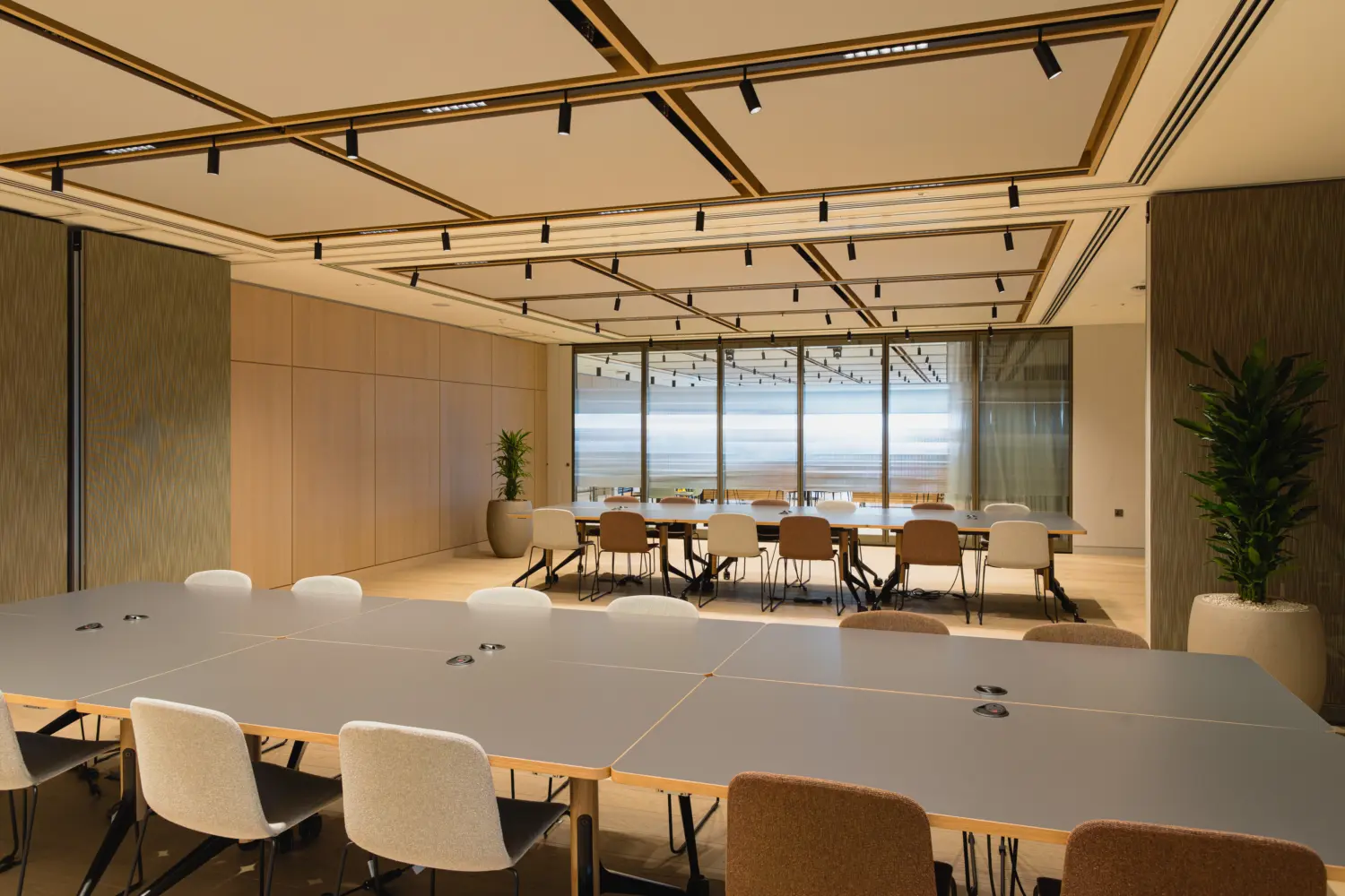 Flexible training and conference room with modular tables and a retractable wall dividing the two main tables. The space features light wood wall panelling, large glazed partitions, integrated ceiling lighting with black track spotlights and a mix of upholstered chairs in neutral tones. Potted plants and natural finishes create a bright, contemporary environment for meetings and collaborative work.
