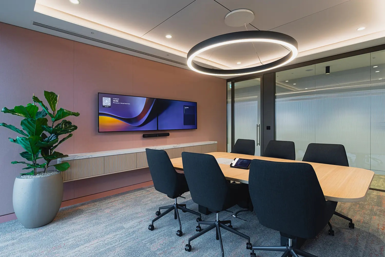 Modern meeting room with a light wood conference table surrounded by black task chairs. A large wall-mounted display screen is set against a muted pink feature wall, with a circular pendant light suspended above the table. Glass partitions, soft carpeting and a large potted plant create a bright, contemporary workspace.