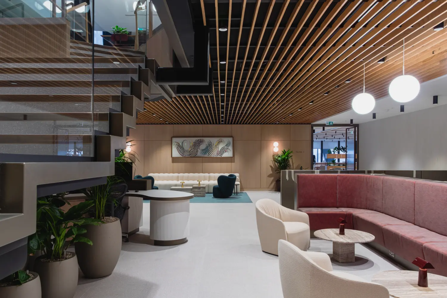 Contemporary office lounge and reception space featuring a timber slatted ceiling, suspended globe lights and a staircase with glass balustrades. Curved pink banquette seating, upholstered lounge chairs and circular tables create informal meeting areas, while planting and light wood finishes add warmth throughout the interior.