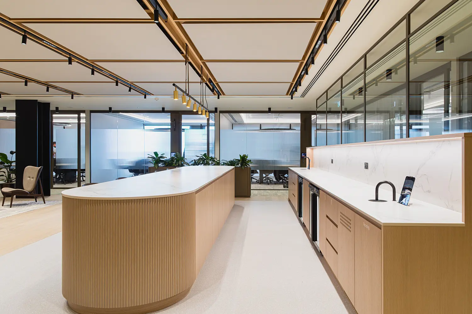 Modern office kitchenette and collaboration hub featuring a large curved island with timber fluted detailing and white stone worktops. Pendant lighting hangs above the island, while built-in cabinetry, sinks and integrated appliances line the rear counter. Glass meeting rooms, indoor planting and a decorative ceiling with integrated track lighting create a bright, contemporary workplace designed for informal gatherings and collaboration.
