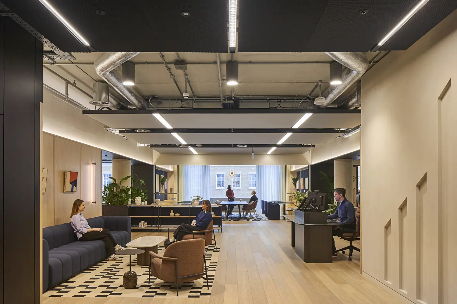 ￼￼Modern office lounge and reception area with light wood flooring, dark ceiling panels, exposed services and linear lighting. A curved blue sofa, armchairs and coffee tables sit on a patterned rug in the foreground, while several people work and meet throughout the open-plan space. Large windows, indoor planting and neutral finishes create a bright, contemporary interior.