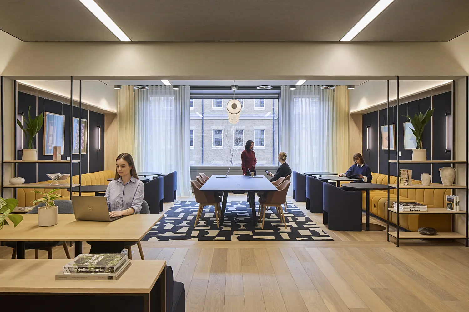 Contemporary office workspace with light timber flooring, built-in booth seating, meeting tables and open shelving. Several people are working throughout the space, including at desks and around a central collaboration table. Dark wall panels, indoor plants, framed artwork and large windows with sheer curtains create a bright, modern environment with a mix of focused work and informal meeting areas.