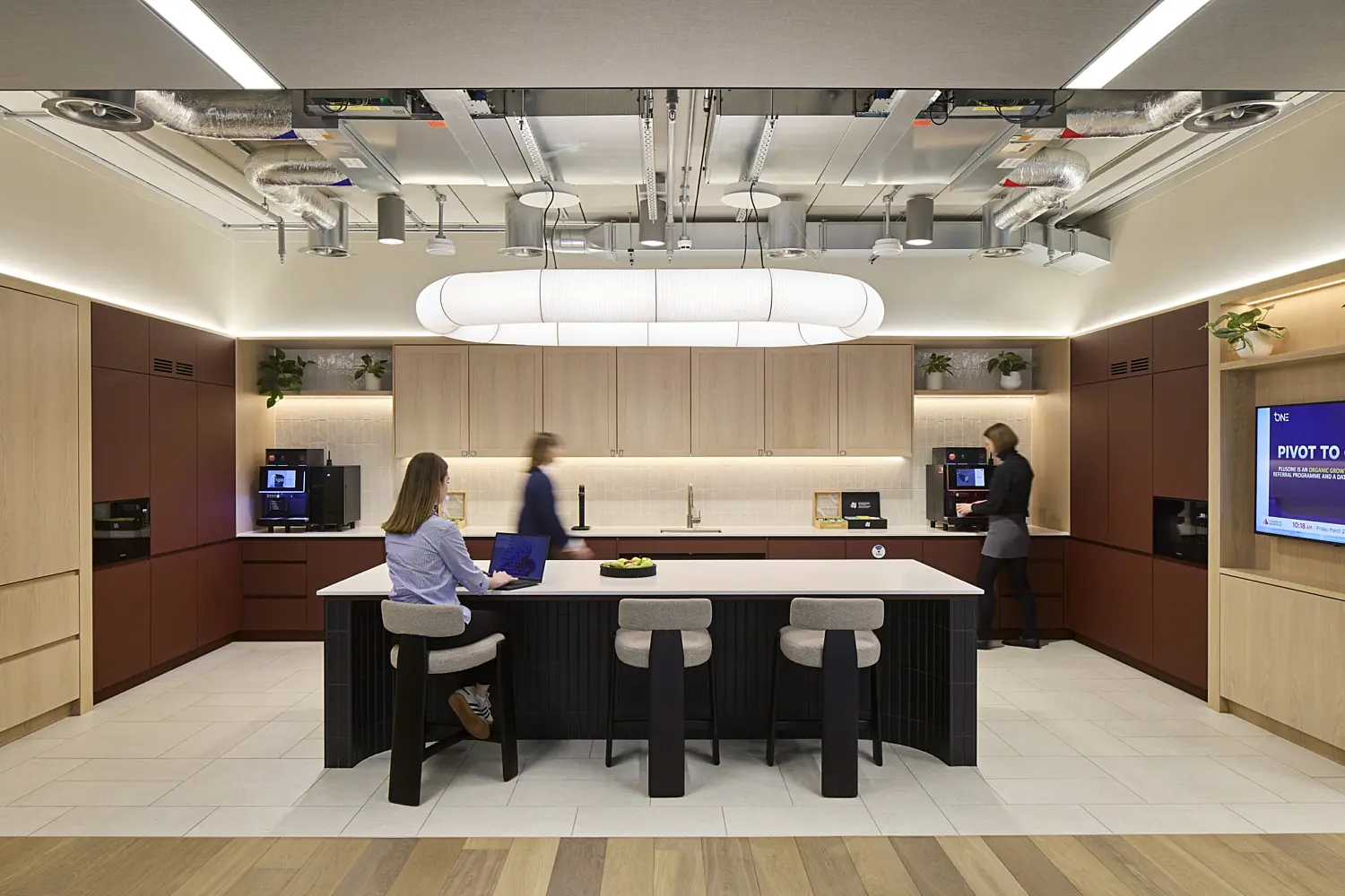 Modern office kitchenette and social hub with a large central island, bar stools and integrated kitchen facilities. Several people are using the space, with one seated at the island working on a laptop while others prepare refreshments. Light timber cabinetry, deep red feature units, indoor planting and exposed ceiling services combine with feature lighting to create a contemporary workplace environment designed for informal meetings, breaks and collaboration.