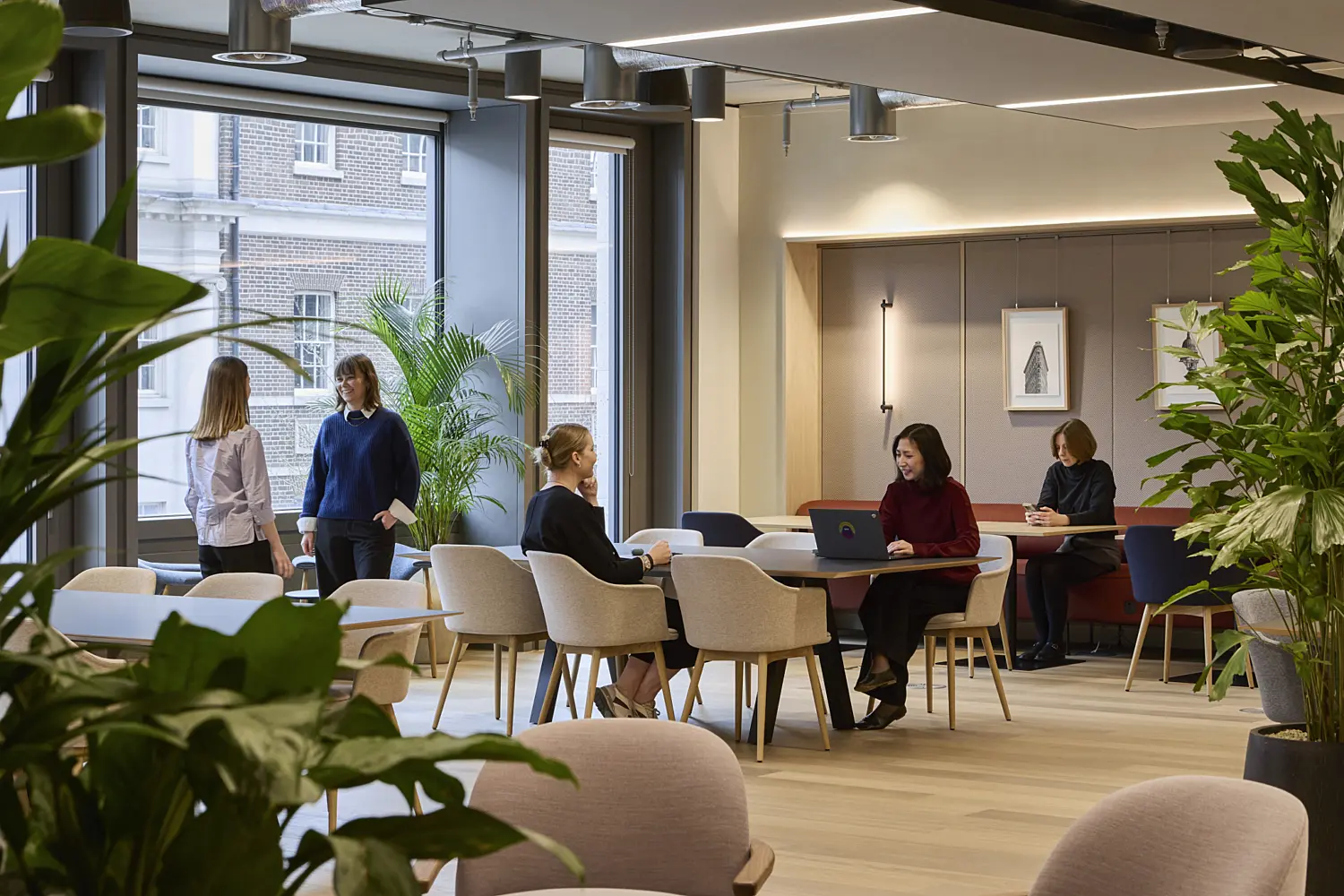 Contemporary office breakout and collaboration space with meeting tables, lounge seating and abundant indoor planting. Several people are working, meeting and conversing throughout the open area, which features light timber flooring, large windows and a mix of soft seating and café-style furniture. Exposed services, acoustic ceiling panels and warm neutral finishes create a bright, welcoming workplace designed for informal collaboration and focused work.