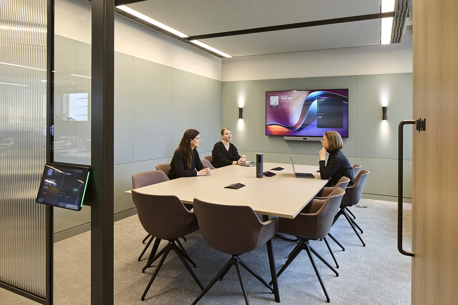 Contemporary meeting room with a large central conference table surrounded by upholstered chairs. Three people are seated around the table, which includes integrated technology for video conferencing and presentations. Soft green wall panels, feature wall lights and a large display screen create a calm, modern setting, while glazed partitions and acoustic ceiling panels help provide privacy and comfort for meetings.