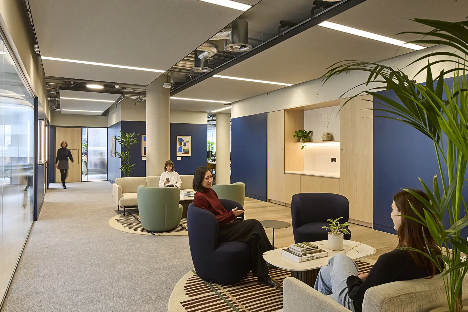 Contemporary office breakout area with lounge seating, circular rugs and coffee tables set within an open circulation space. Several people are seated in informal meeting areas while another walks through the corridor. Blue feature walls, light timber joinery, indoor plants and acoustic ceiling panels with integrated lighting create a bright, modern workplace designed for collaboration and relaxation.