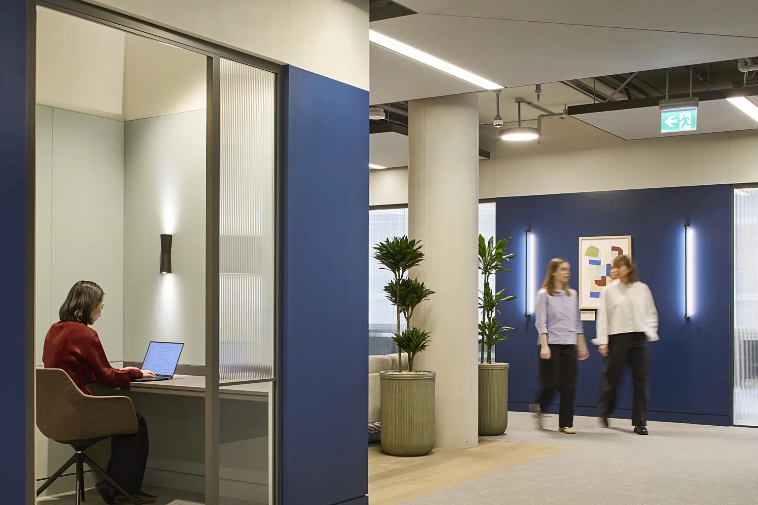 Modern office corridor with a quiet focus booth on one side, where a person is seated at a desk using a laptop. The space features blue and neutral wall finishes, large planters, exposed services and acoustic ceiling panels with integrated lighting. In the background, two people walk through the open circulation area, with artwork and feature wall lights adding colour to the contemporary workplace interior.