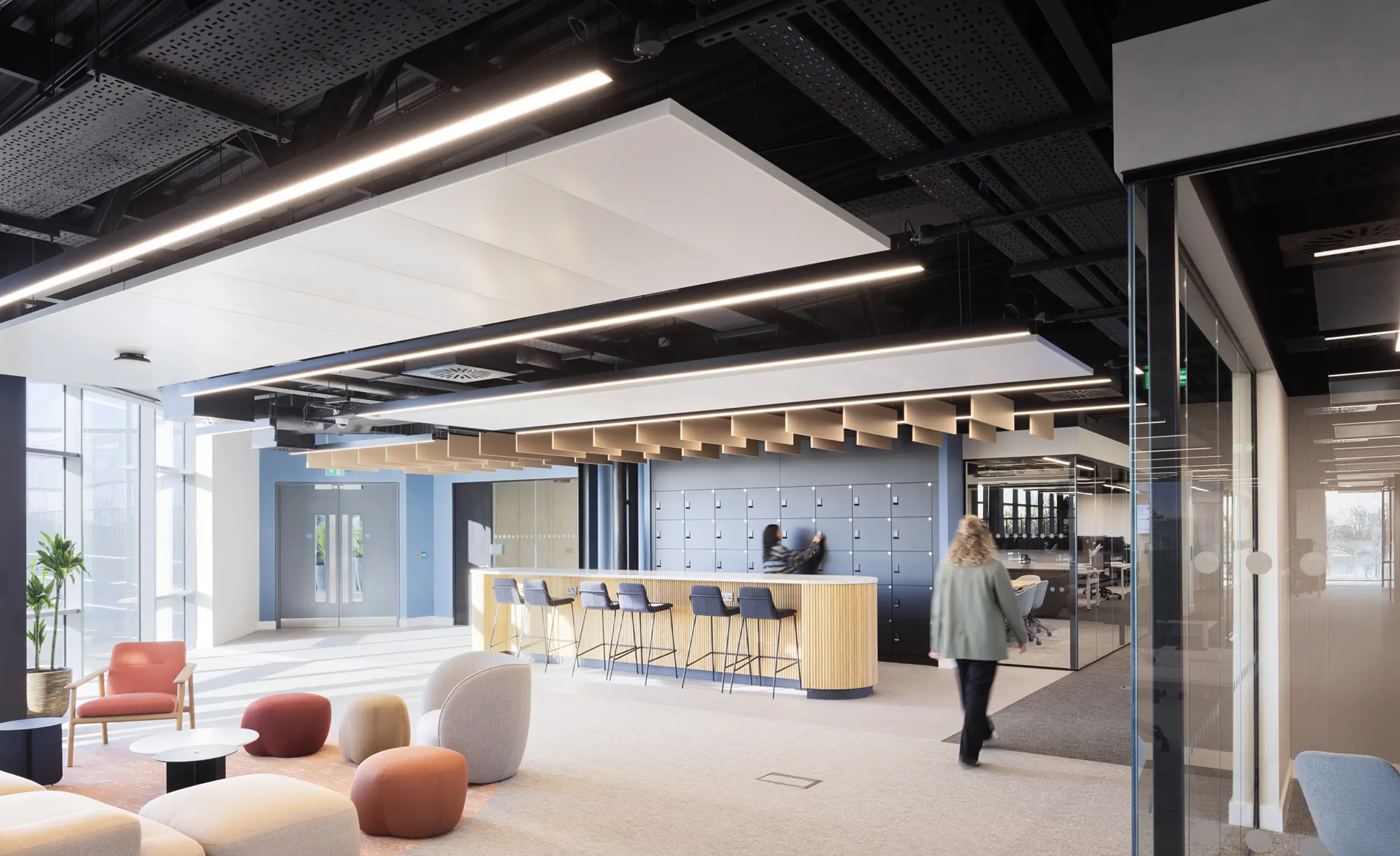 A bright open plan office with contemporary furniture including soft seating in muted colours at the front, a long timber island with bar stools in the centre and a dark feature wall with lockers behind it. The ceiling shows exposed services with suspended lighting panels. Floor to ceiling windows on the left bring in natural light. A person is walking across the space toward a glass meeting room on the right, and another person is seated at the island.