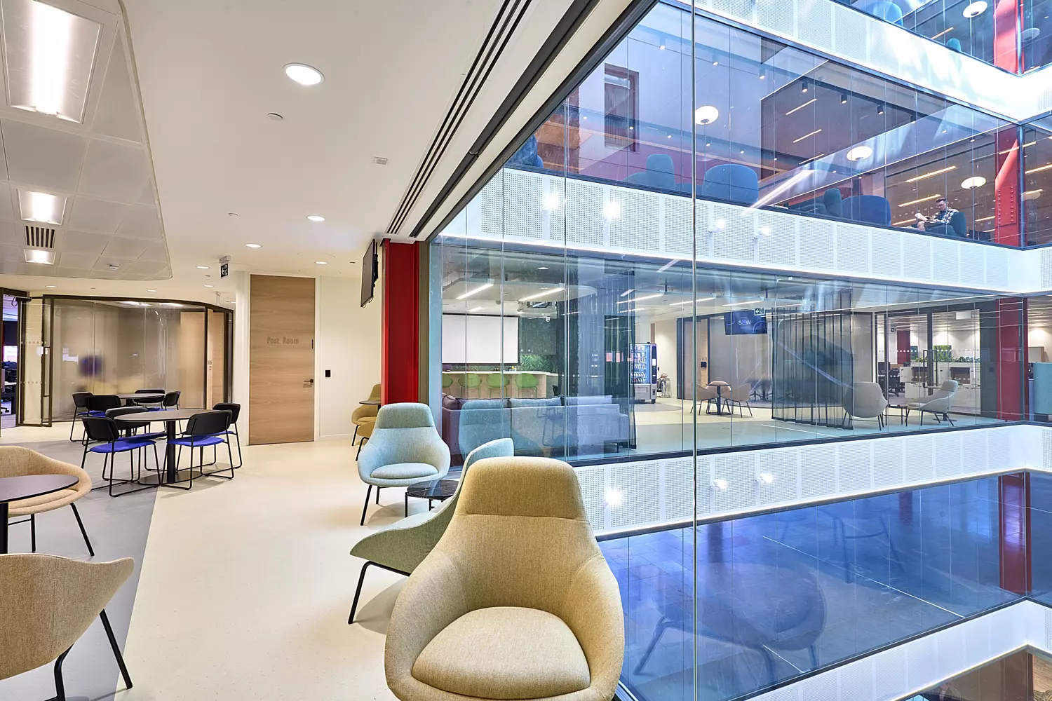 Contemporary office breakout space with soft seating in neutral tones positioned near a full-height glass wall overlooking an open atrium. The design features clean lines, light flooring, and modern furniture with curved backs. Visible across the atrium are multiple floors with collaborative zones, colourful accents, and glass balustrades, creating a bright and open environment that encourages connectivity and transparency.