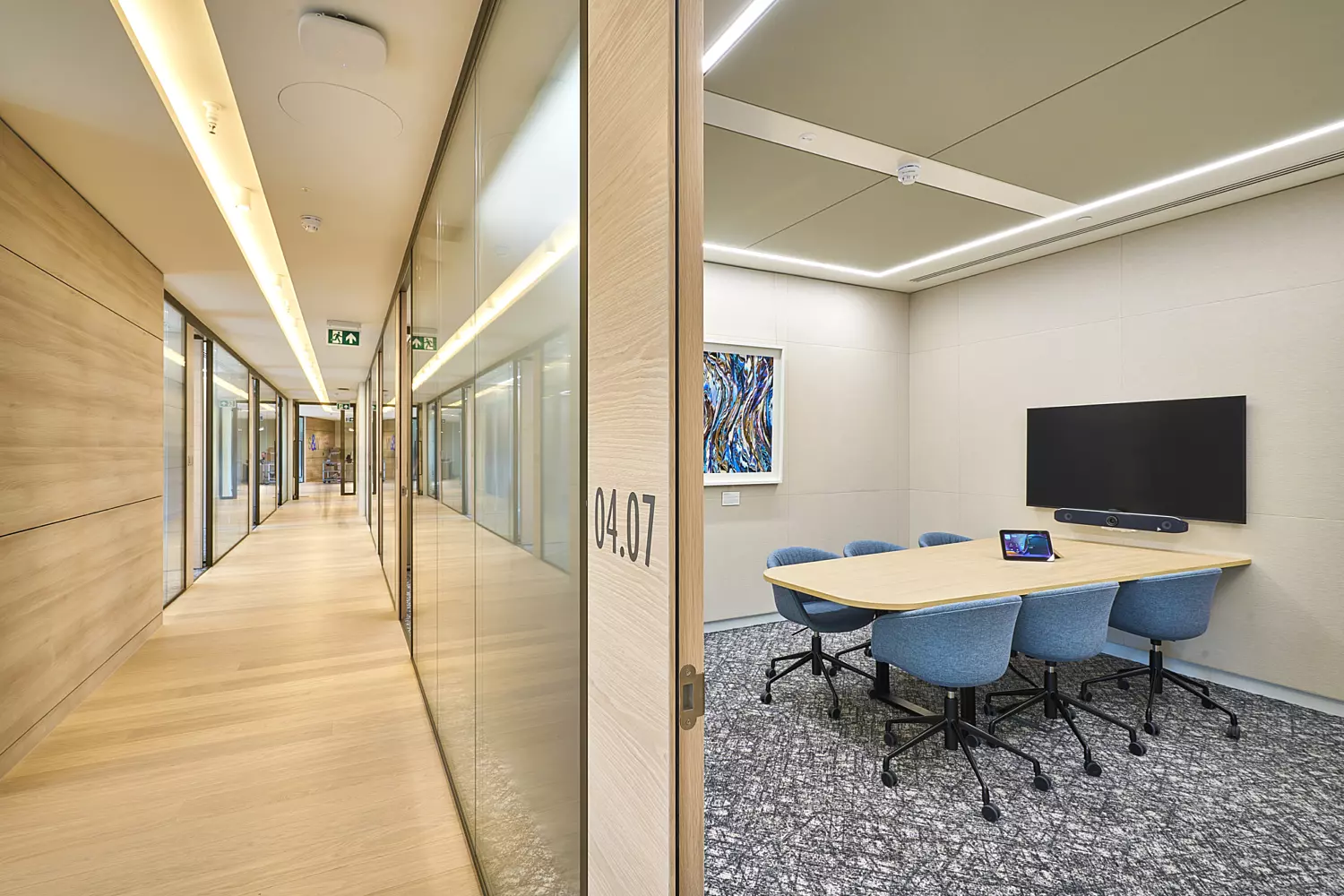 Modern meeting room with a rectangular wooden table and six blue upholstered chairs on a patterned grey carpet. The space includes a wall-mounted display screen and integrated video conferencing equipment. Neutral walls and a minimalist ceiling with recessed linear lighting create a clean, contemporary look. Adjacent to the room is a bright corridor with timber wall panels, glass partitions, and light wood flooring, adding warmth and transparency to the office design.