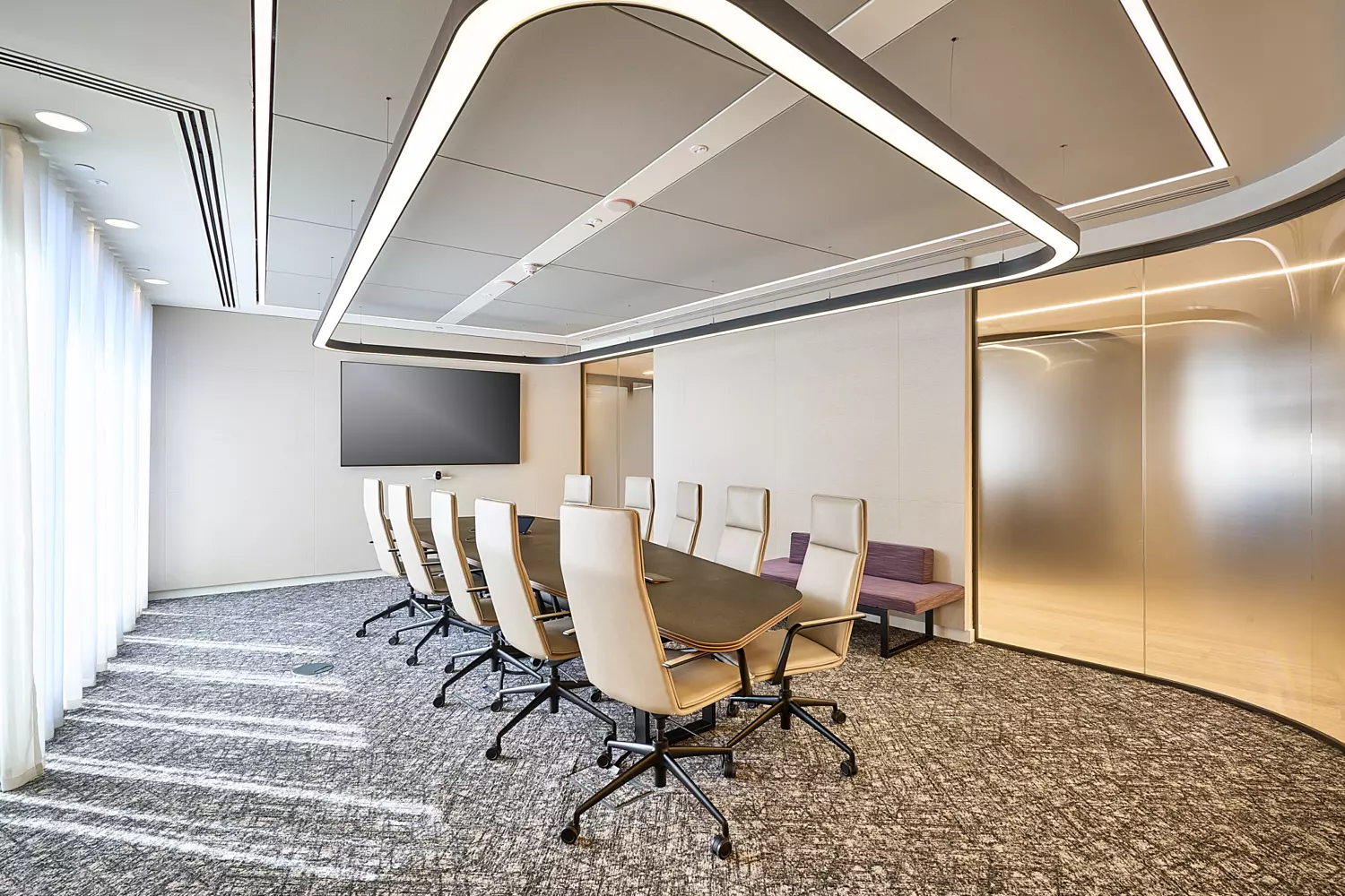 Modern boardroom featuring a long rectangular meeting table with high-back chairs arranged around it. The space has a patterned grey carpet, light neutral walls, and a large wall-mounted display screen. A standout feature is the ceiling design with integrated LED lighting in geometric shapes, creating a sleek and contemporary look. Frosted glass partitions add privacy while maintaining an open feel, and sheer curtains allow natural light to filter through.