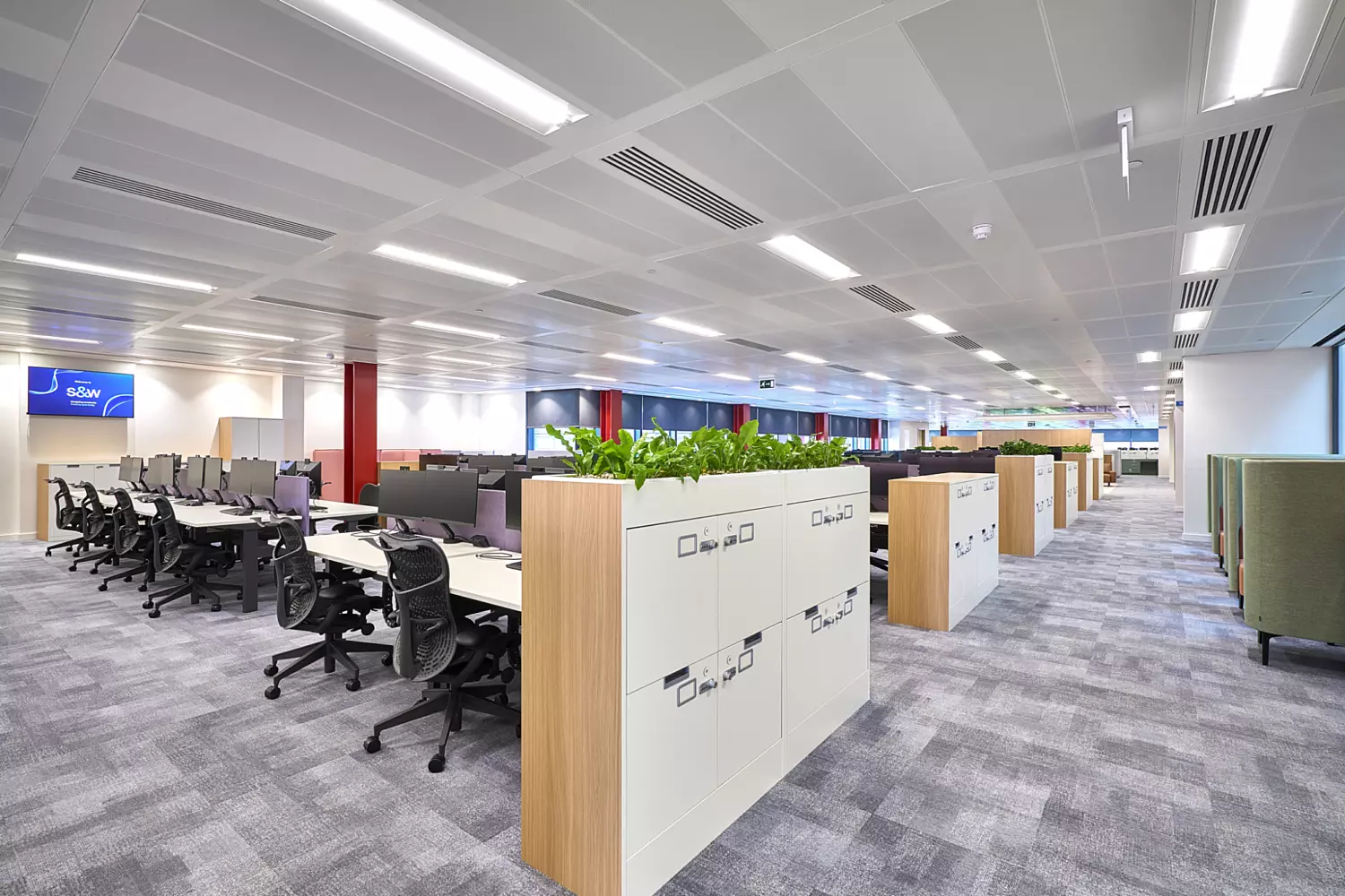 Large open-plan office with rows of workstations featuring ergonomic chairs and dual monitors. The space includes light grey patterned carpet, white acoustic ceiling panels with integrated lighting, and bold red columns for visual contrast. Storage units with built-in planters add greenery and create natural zoning. The design promotes flexibility and collaboration in a bright, modern environment.