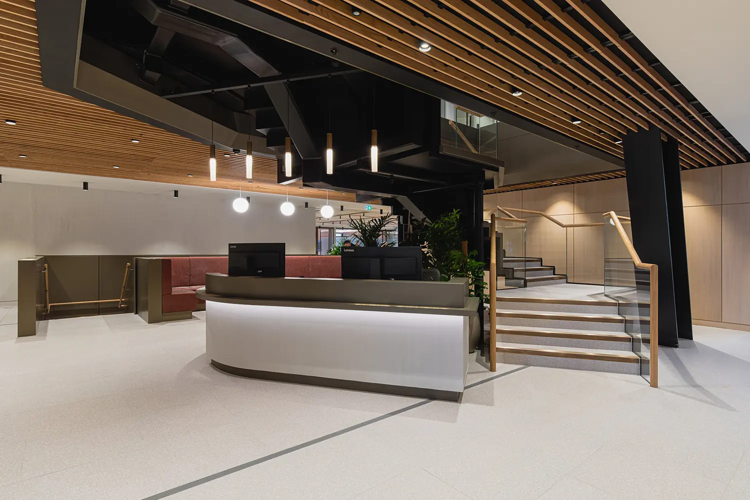 Modern office reception area with a curved white reception desk, light stone flooring and a striking timber slatted ceiling. Black structural features and pendant lights create contrast, while staircases with glass balustrades lead to upper levels. Built-in seating and indoor planting add warmth to the contemporary lobby space.