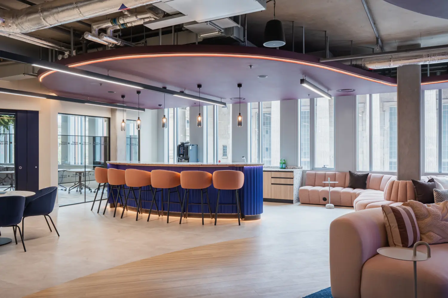 Modern office breakout space with a curved blue bar and peach-upholstered stools beneath a suspended ceiling feature with integrated lighting. Large windows bring in natural light, while soft seating areas with curved sofas and small tables create informal meeting and collaboration spaces. Exposed services and contemporary finishes give the interior a stylish, open-plan feel.