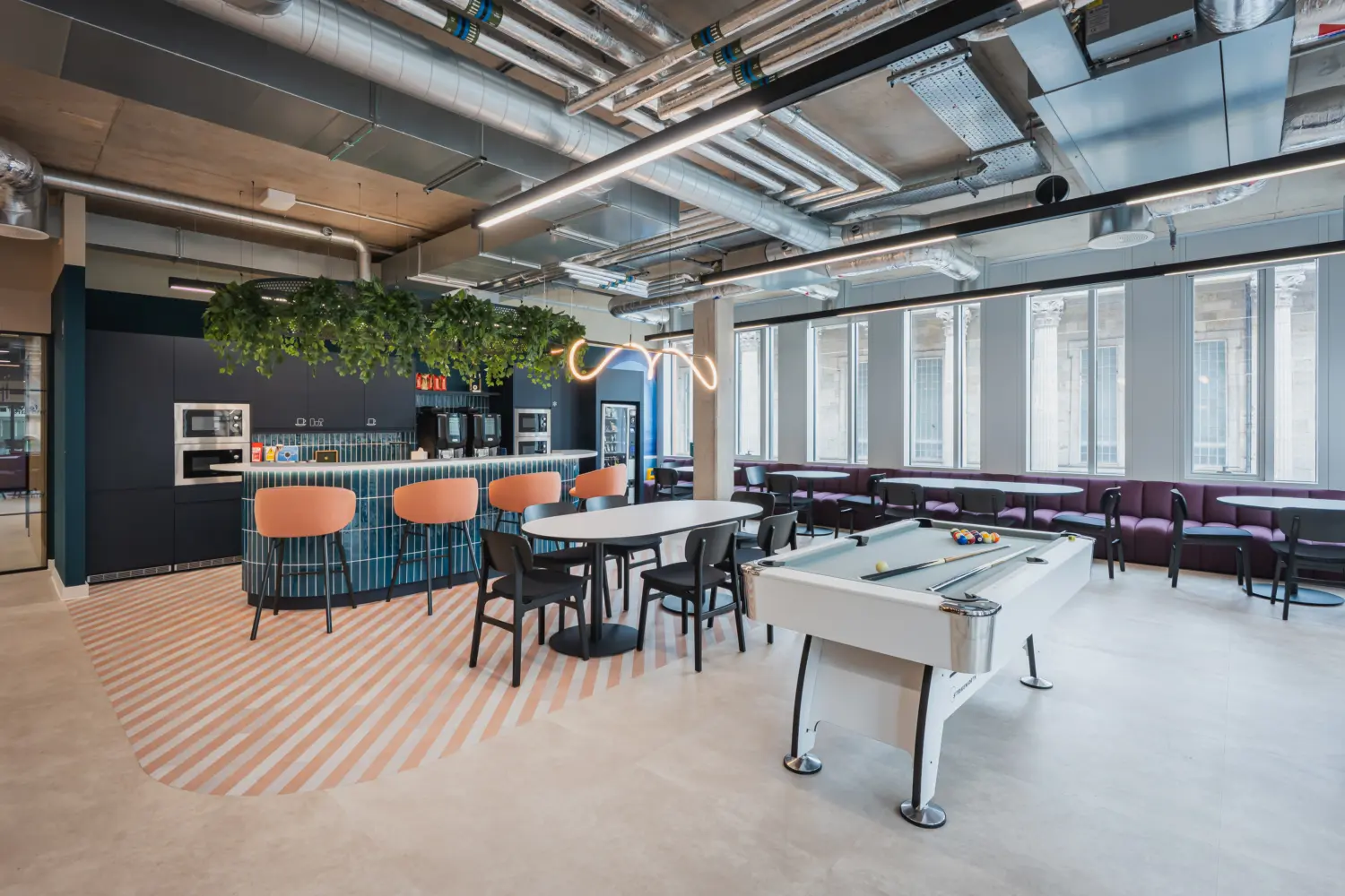 Contemporary office café and social space featuring a kitchenette with teal panelling, peach bar stools and overhead planting. Round dining tables and booth seating sit alongside a pool table, creating a relaxed area for informal meetings and breaks. Large windows provide natural daylight, while exposed services and modern lighting contribute to an industrial-inspired workplace design.