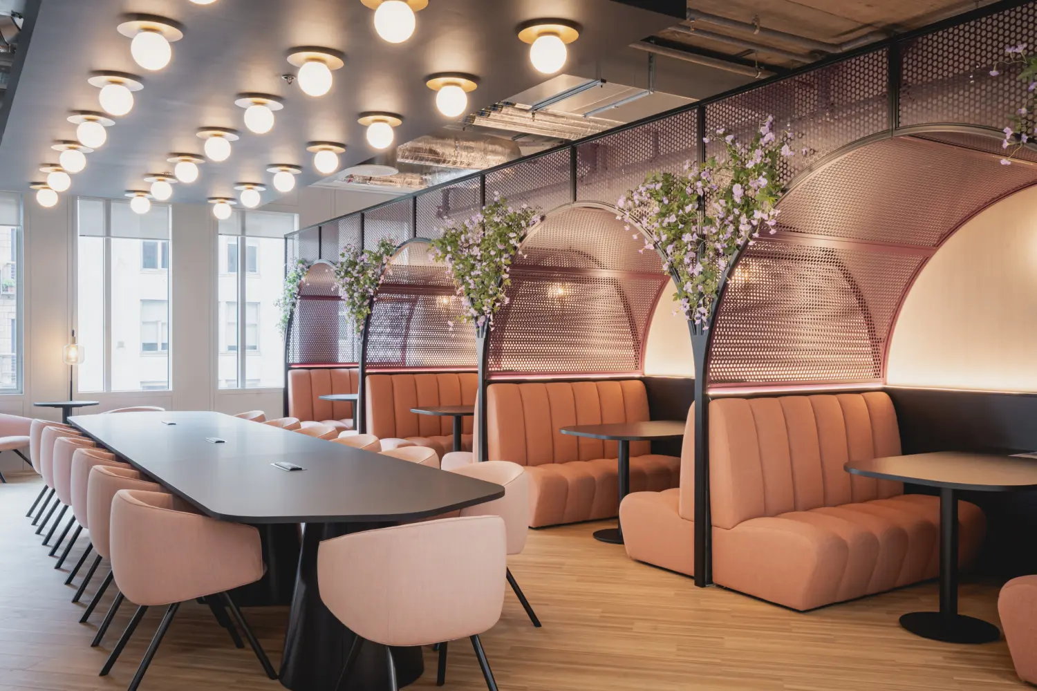 Modern office collaboration area featuring a long communal table surrounded by upholstered chairs and a series of semi-enclosed booth seating areas with curved metal screens and integrated planting. Soft pink and copper tones, warm lighting and decorative floral features create a welcoming atmosphere. Large windows provide natural daylight, while exposed ceiling services and contemporary finishes give the space a modern workplace