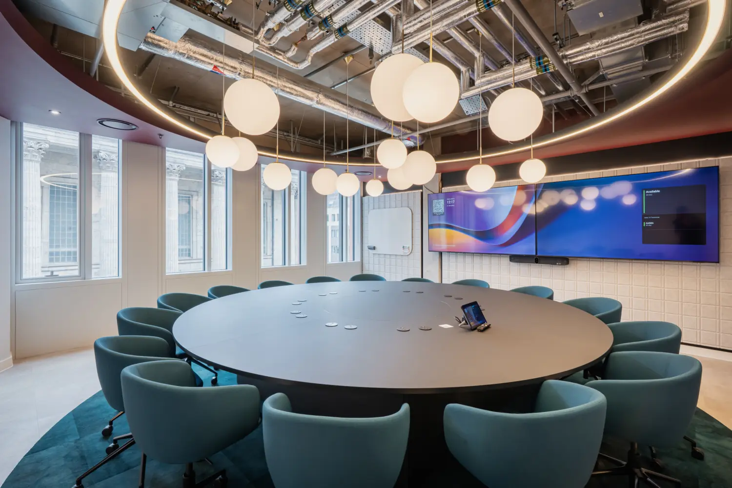 Modern meeting room featuring a large circular conference table surrounded by teal upholstered chairs. Pendant globe lights hang beneath an illuminated circular ceiling feature, while dual wall-mounted display screens and a whiteboard provide presentation facilities. Floor-to-ceiling windows bring in natural daylight, and exposed ceiling services combined with contemporary finishes create a stylish, collaborative workspace.