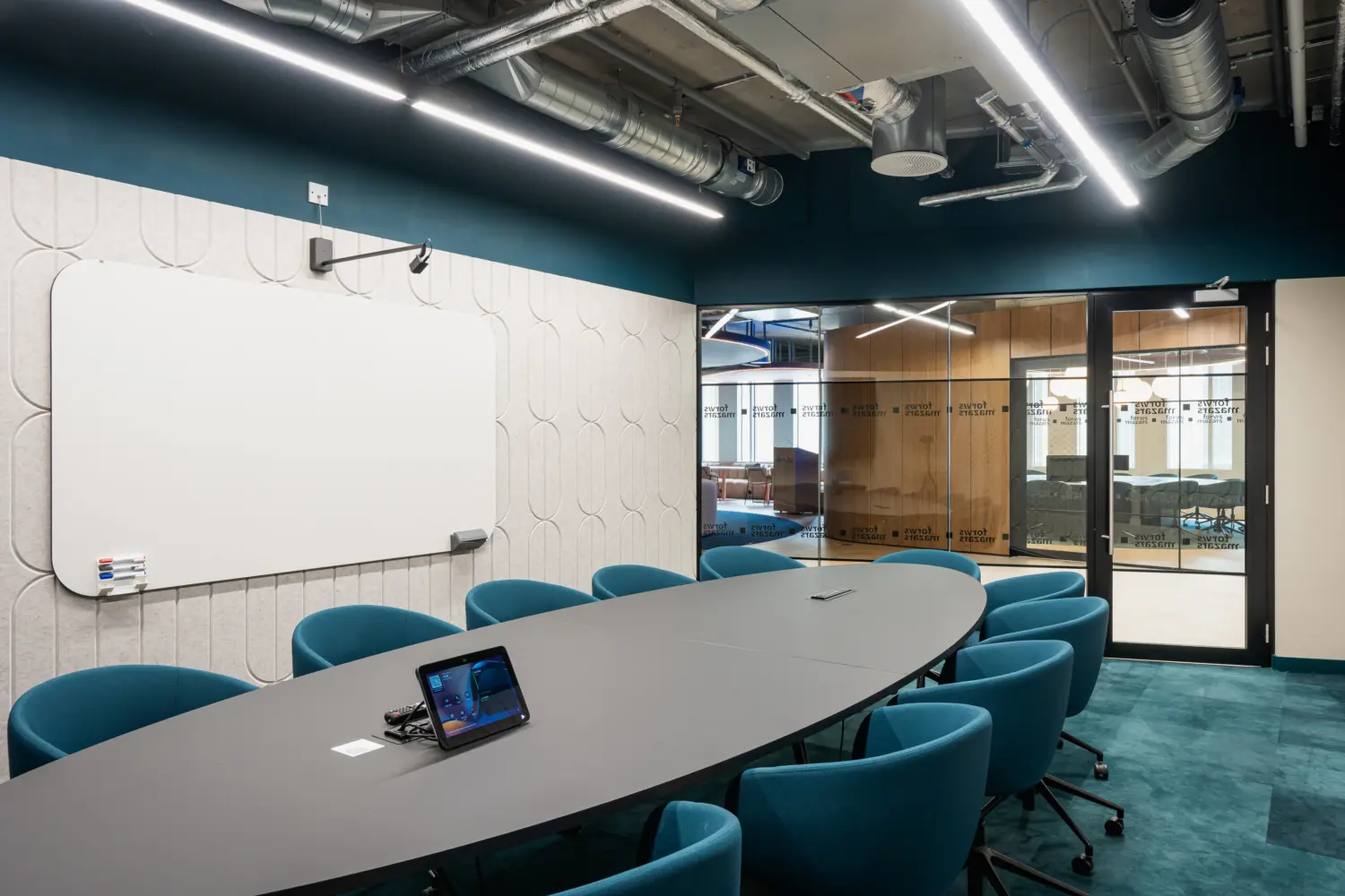 Contemporary meeting room featuring an oval conference table surrounded by teal upholstered chairs. A large wall-mounted whiteboard provides space for presentations and collaboration, while integrated meeting technology sits on the table. Glass partitions and timber detailing connect the room to the surrounding office, and exposed ceiling services with suspended linear lighting create a modern, industrial-inspired workplace aesthetic.