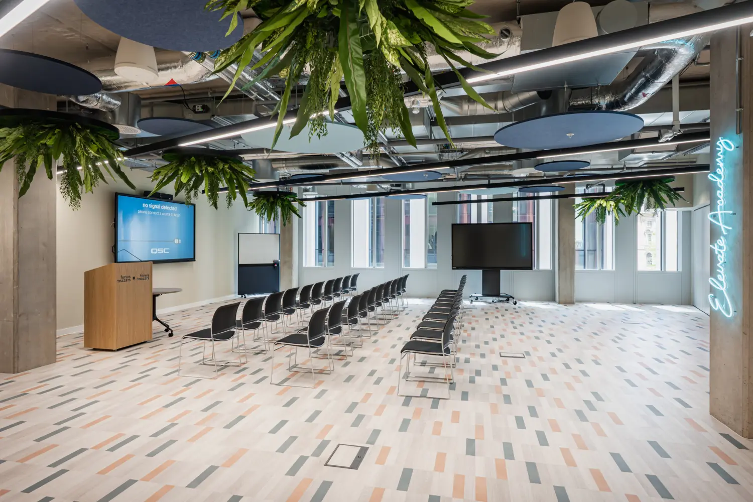 Large multi-purpose event space within a contemporary office, featuring rows of chairs facing presentation screens and a lectern. Suspended planting and circular acoustic panels are integrated throughout the exposed ceiling, while floor-to-ceiling windows provide natural daylight. Patterned flooring, concrete columns and modern AV technology create a flexible environment for town halls, presentations and team gatherings.