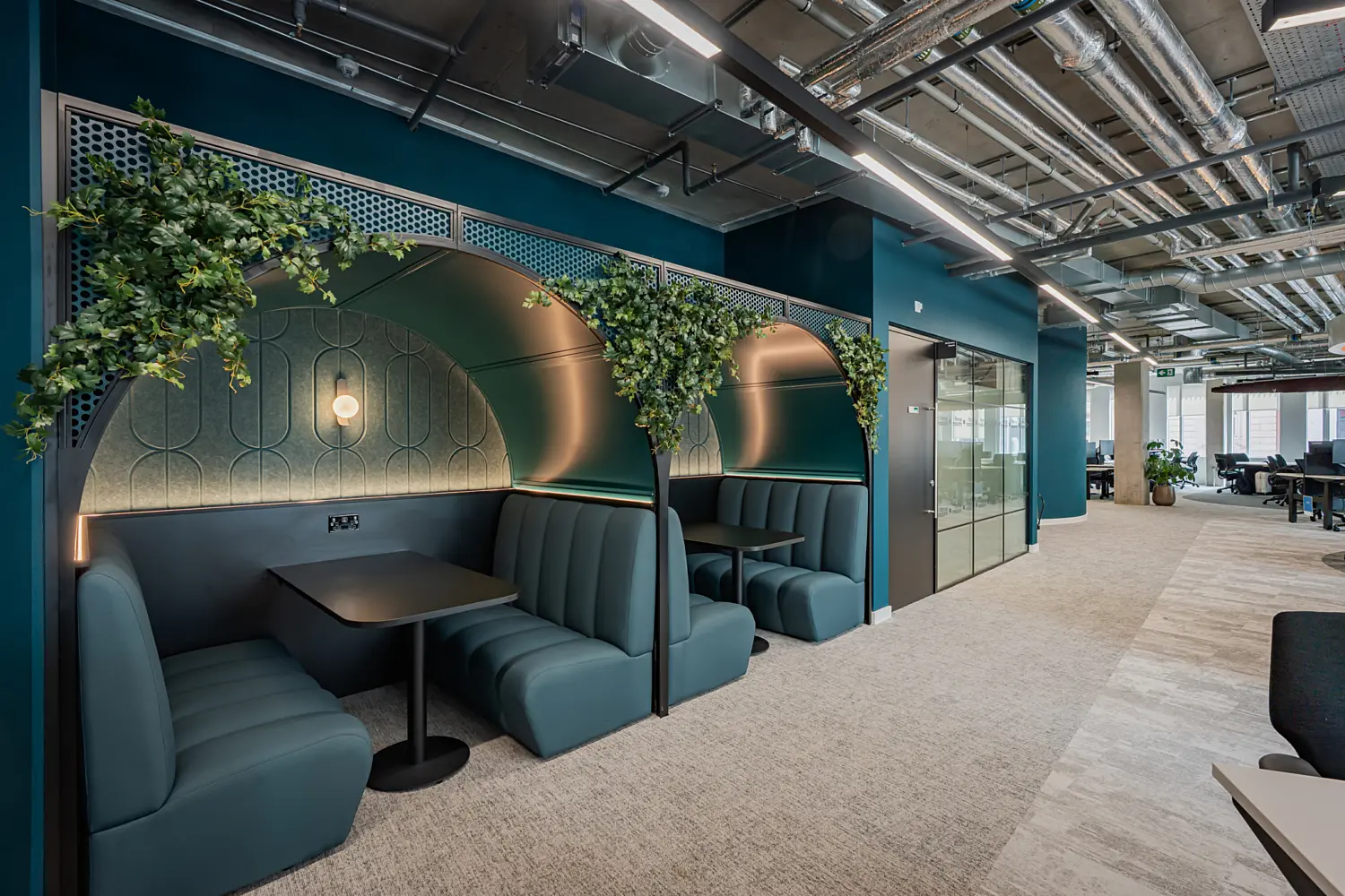 Contemporary office workspace featuring a row of semi-enclosed booth seating areas with teal upholstered benches, integrated lighting and decorative planting. The booths provide informal spaces for meetings and focused work, set alongside an open-plan office with desks, glazed meeting rooms and indoor greenery. Exposed ceiling services, linear lighting and rich blue-green finishes create a modern, industrial-inspired workplace environment.