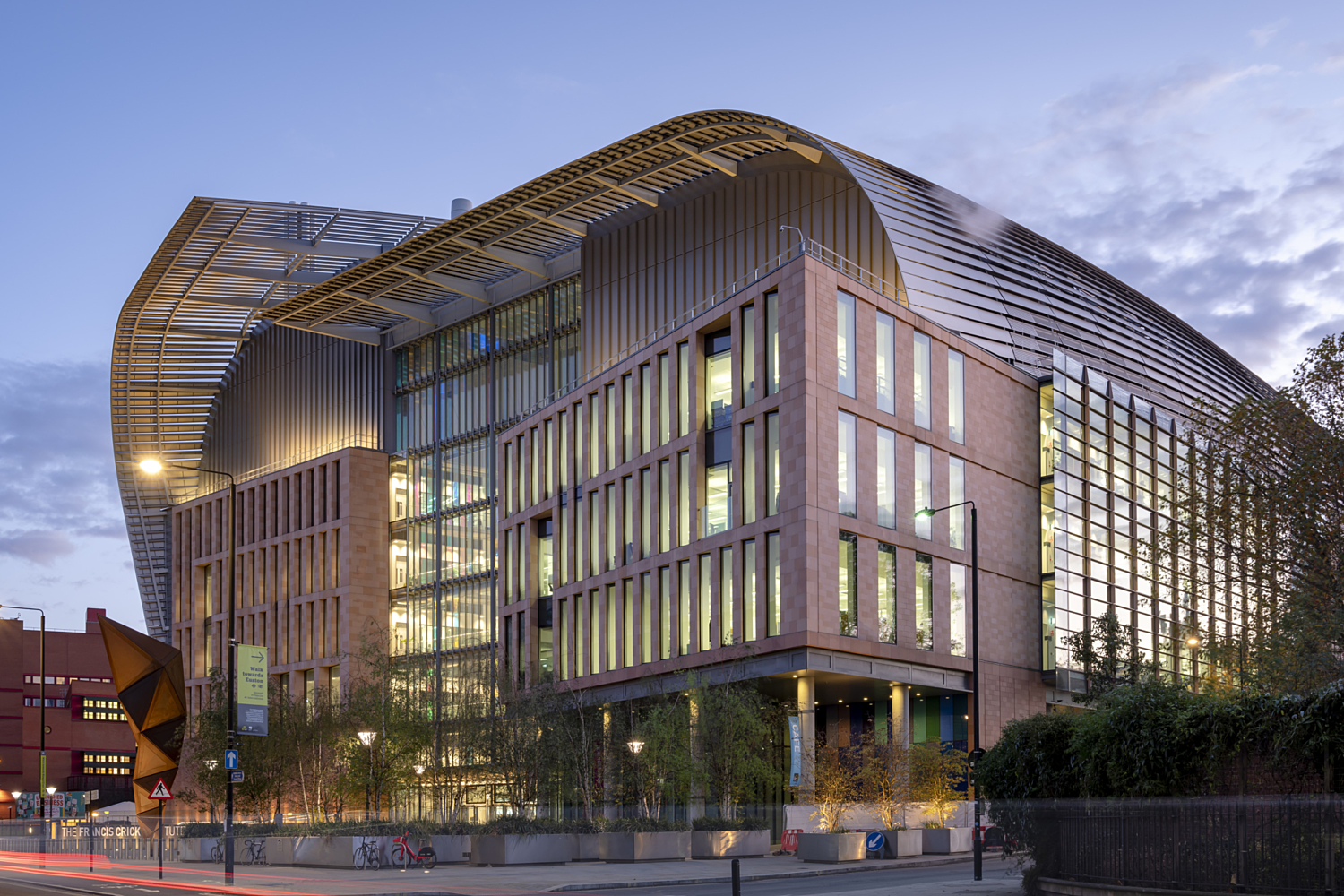 Architecturally distinctive building with a curved roof and large glass windows spanning multiple storeys. The exterior combines glass and brick materials, creating a visually striking contrast. In the foreground, trees and streetlights line the street, adding context to the urban setting