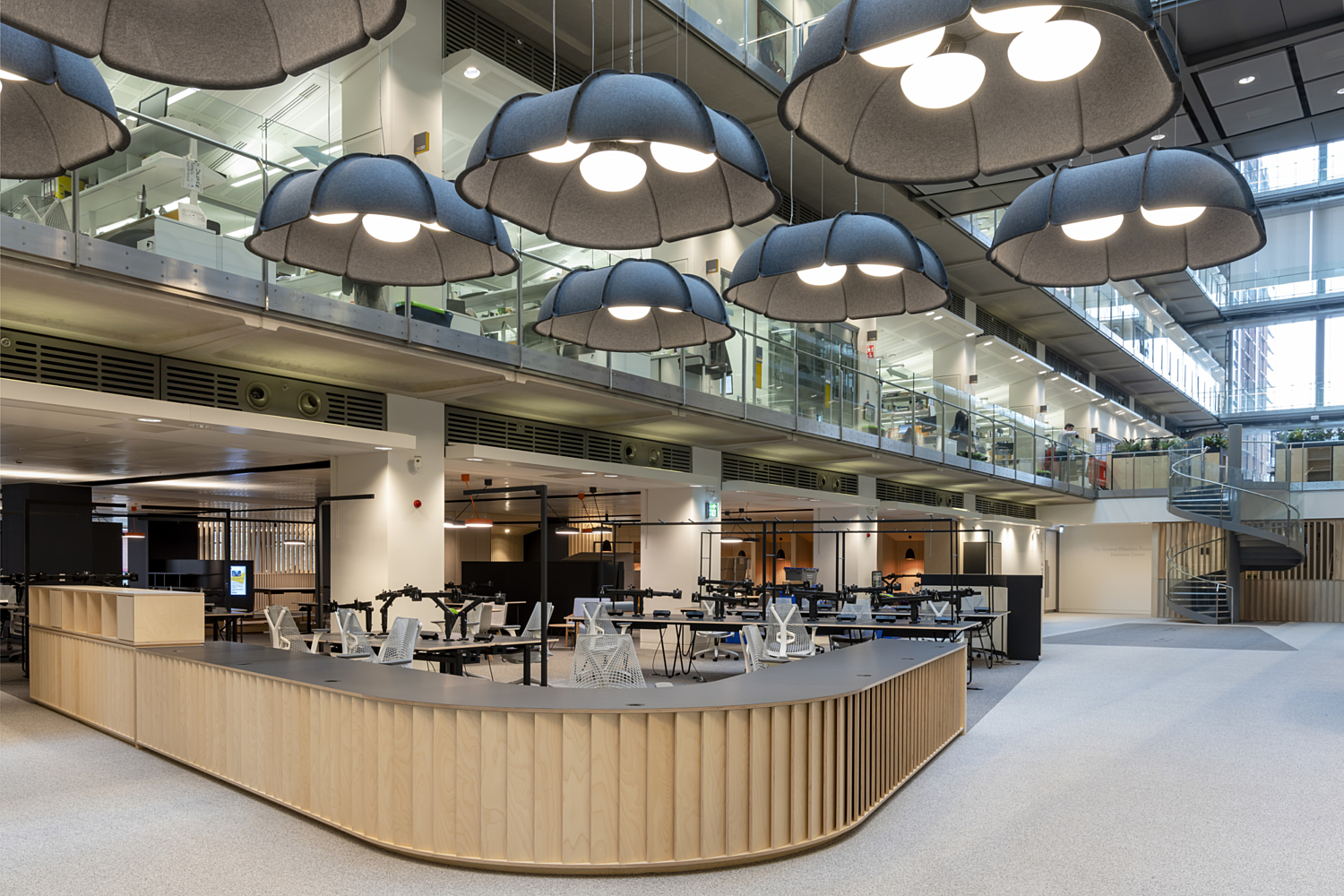 A spacious, modern reception area at the Francis Crick Institute featuring a sleek, curved wooden reception desk with vertical slats and a matte grey counter. Overhead, a cluster of oversized pendant lights with soft, umbrella-like fabric shades add a sculptural element and diffuse lighting. The open-plan layout includes glass-walled upper floors with visible office spaces, exposed concrete beams and clean architectural lines, creating a blend of industrial and contemporary design. Neutral tones and natural materials give the space a calm, professional feel.