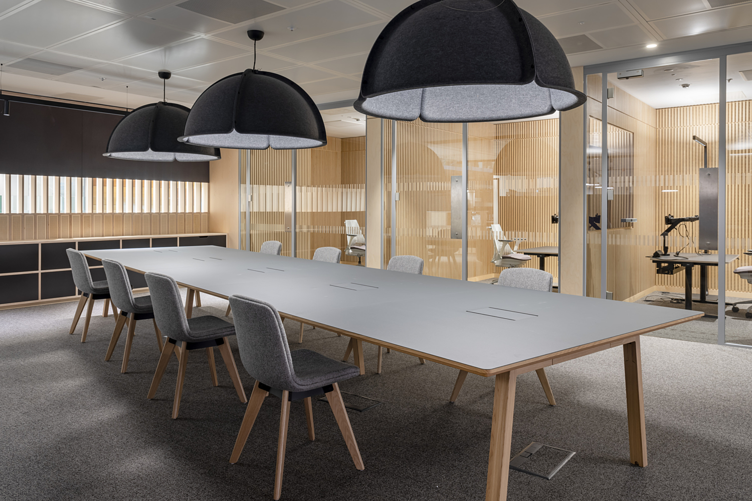 Room with a long rectangular table surrounded by multiple chairs, illuminated by three large dome-shaped pendant lights suspended from the ceiling. The space features a combination of glass and wood walls, with an adjacent office area visible through the glass partition. The overall design is clean and professional, suited for meetings and collaborative work.