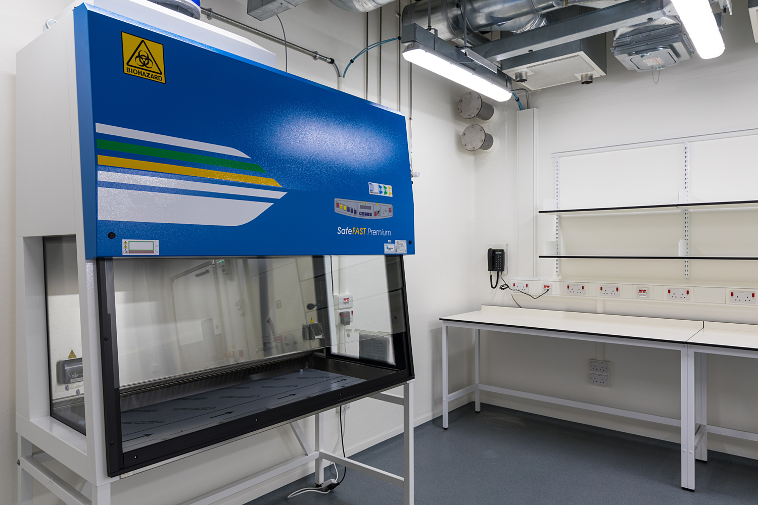 Laboratory room featuring a blue and white fume hood on the left, labelled 'ESCO' and 'Labculture,' with a transparent front panel and integrated control systems. On the right, an empty workbench sits beneath a row of electrical outlets and switches mounted on the wall. The space is illuminated by overhead lighting, with ventilation ducts visible along the ceiling, reflecting a clean and functional laboratory environment.