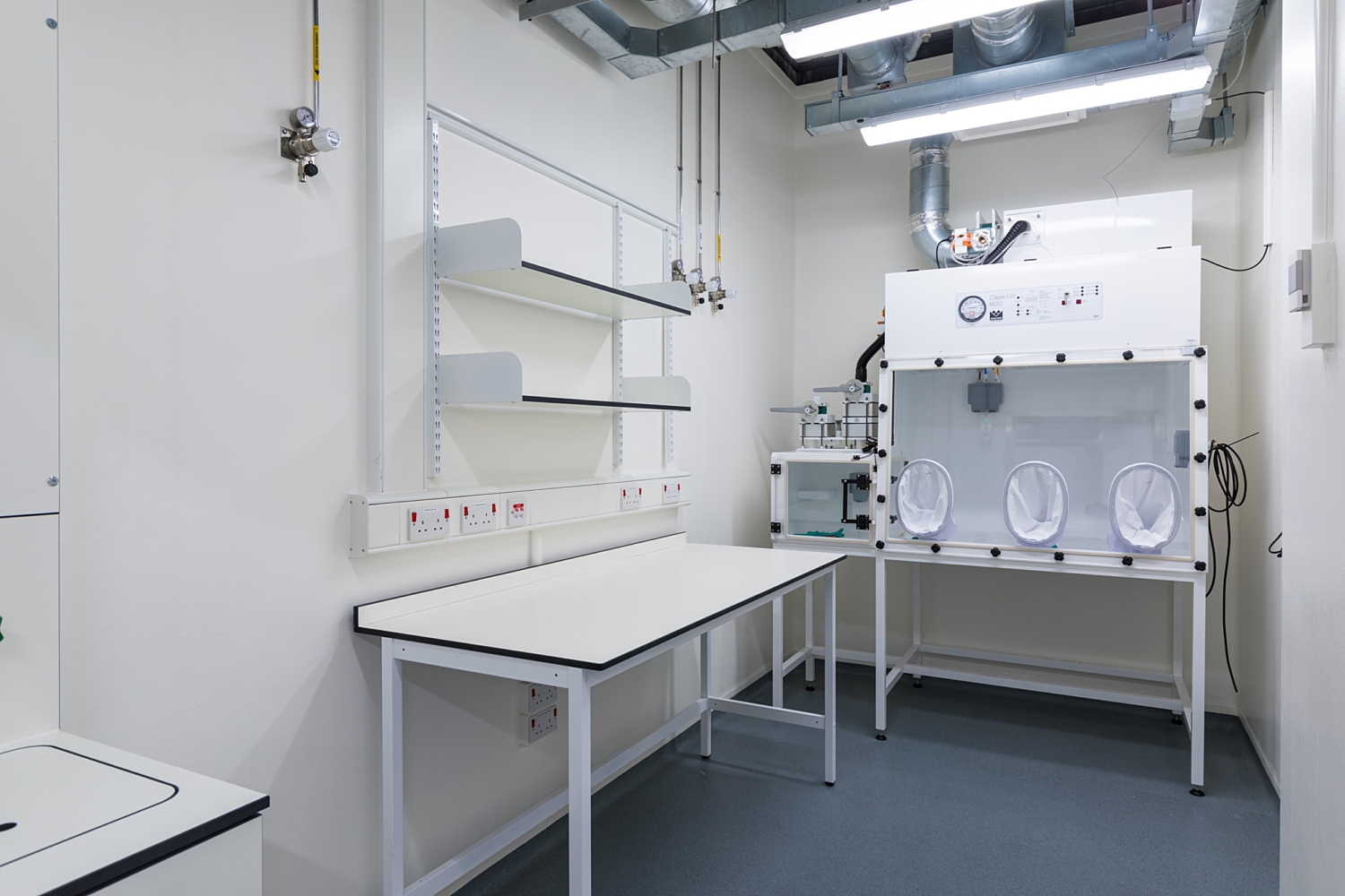 Laboratory space with white walls and grey flooring, designed for controlled scientific work. On the left, a workbench with shelving provides storage and workspace, while on the right, a glove box system with three attached gloves allows for the safe handling of hazardous materials. The room includes electrical outlets, switches, and visible ceiling-mounted pipes and ventilation ducts, all contributing to a functional and safety-focused environment.