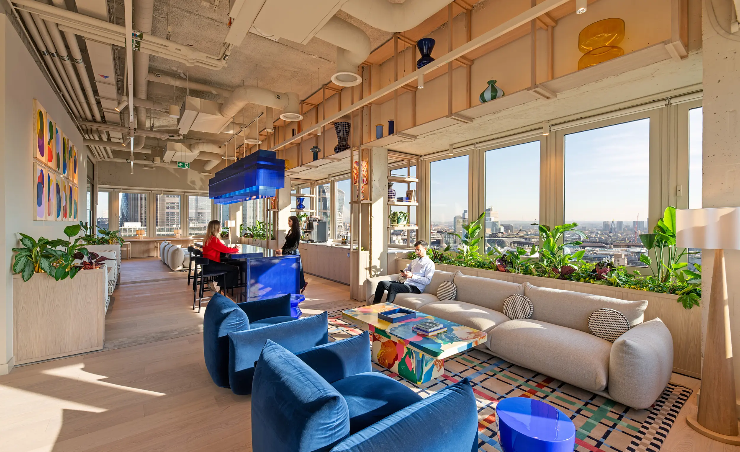 Modern office lounge with floor-to-ceiling windows overlooking the London skyline. A light-coloured sofa and blue upholstered chairs surround a colourful coffee table on a patterned rug. Plants line the windows, while a blue statement pendant light hangs above a central bar where two people are seated. Exposed ceilings, artwork and decorative shelving create a bright, contemporary workspace flooded with natural daylight.