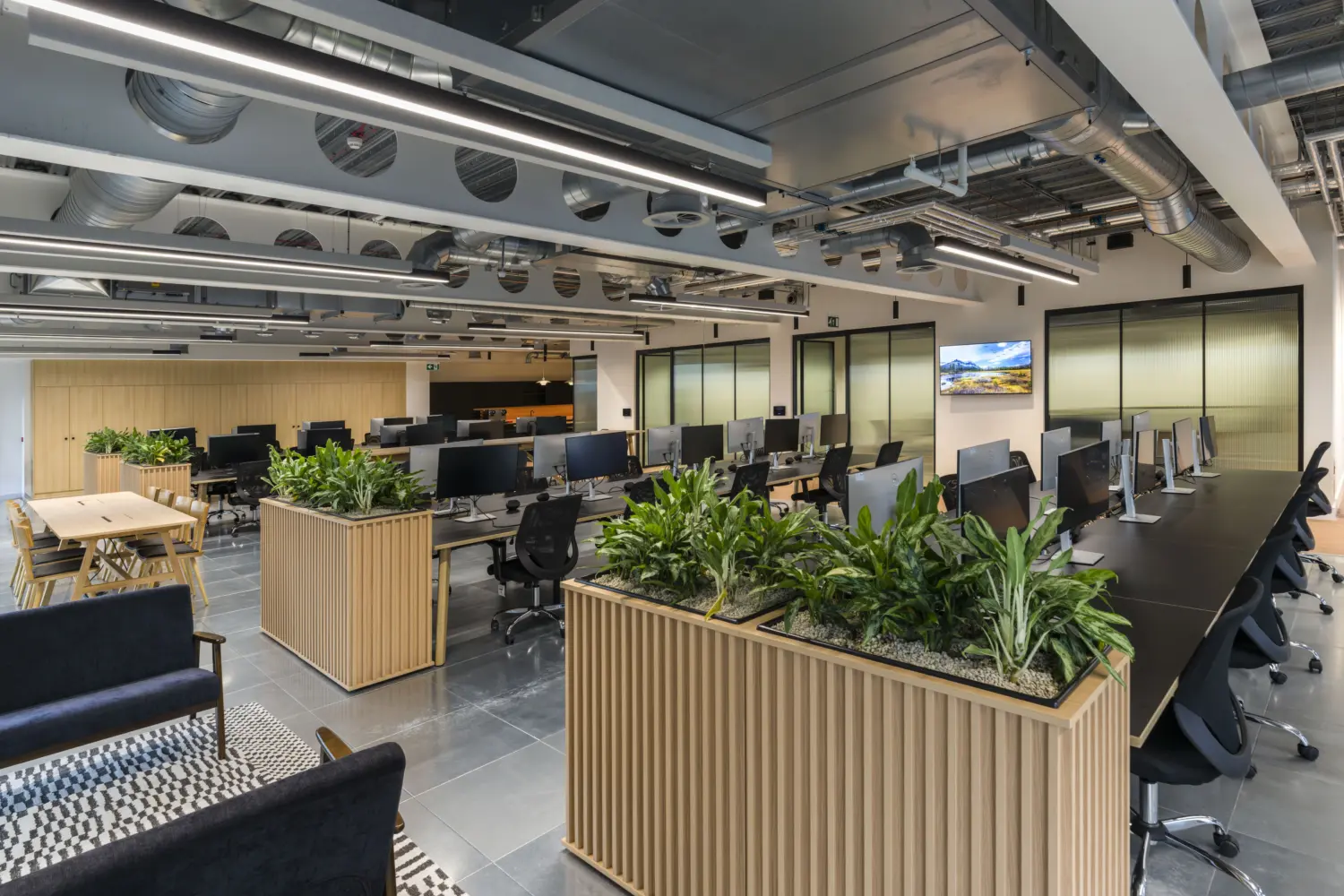 Open-plan office workspace with rows of desks and computer monitors arranged around large timber planters filled with greenery. The interior features exposed ceiling services, suspended linear lighting and glazed meeting rooms along the perimeter. Informal seating and a shared table are positioned in the foreground, while light wood joinery and planting introduce warmth throughout the contemporary office environment.