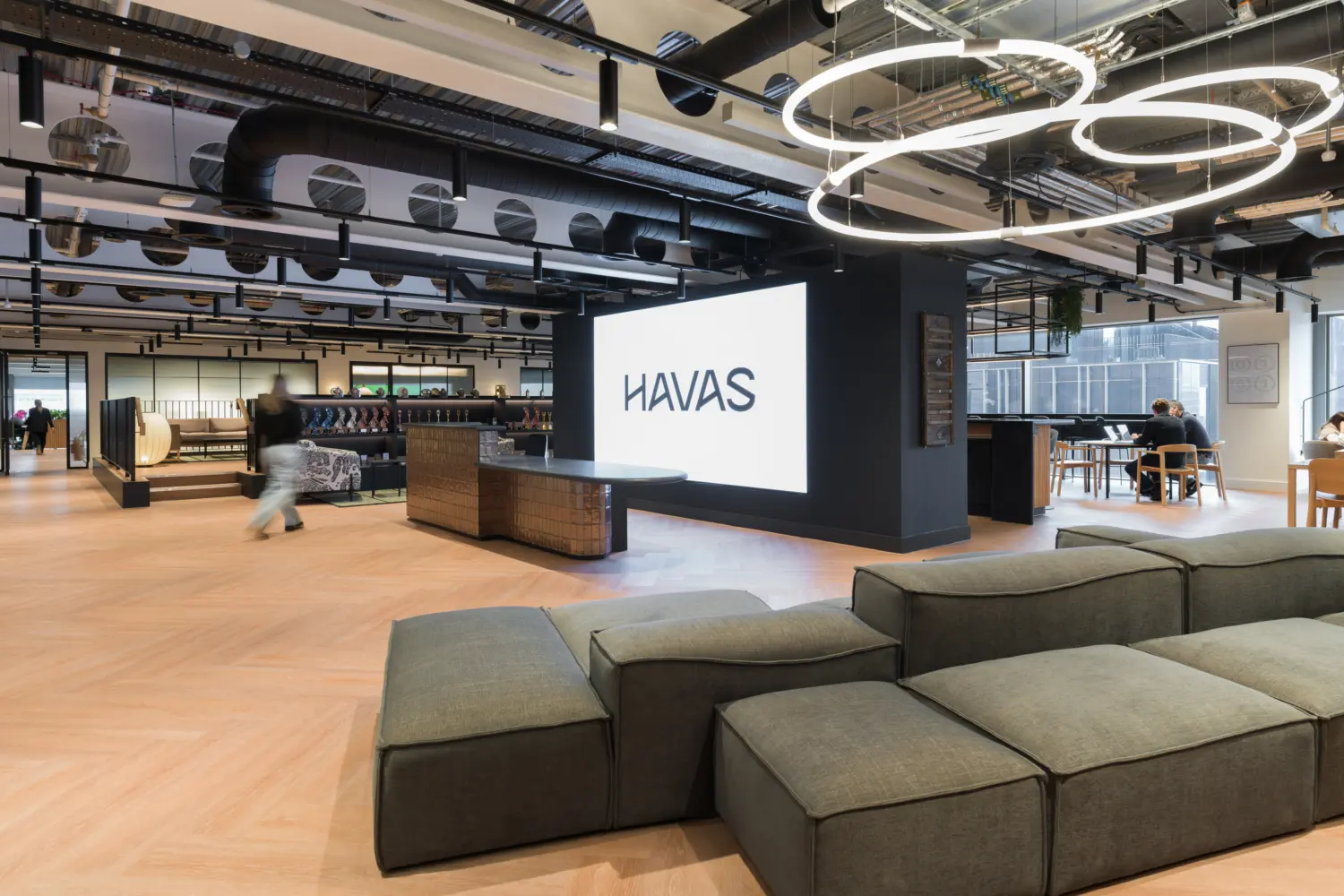 Modern open-plan office lounge with light wood flooring, exposed ceiling services and circular LED light fixtures. A large illuminated HAVAS logo is displayed on a central feature wall beside a curved reception desk. Modular grey seating fills the foreground, while booths, tables and workspaces extend across the bright interior. Several people are visible using the space near floor-to-ceiling windows.