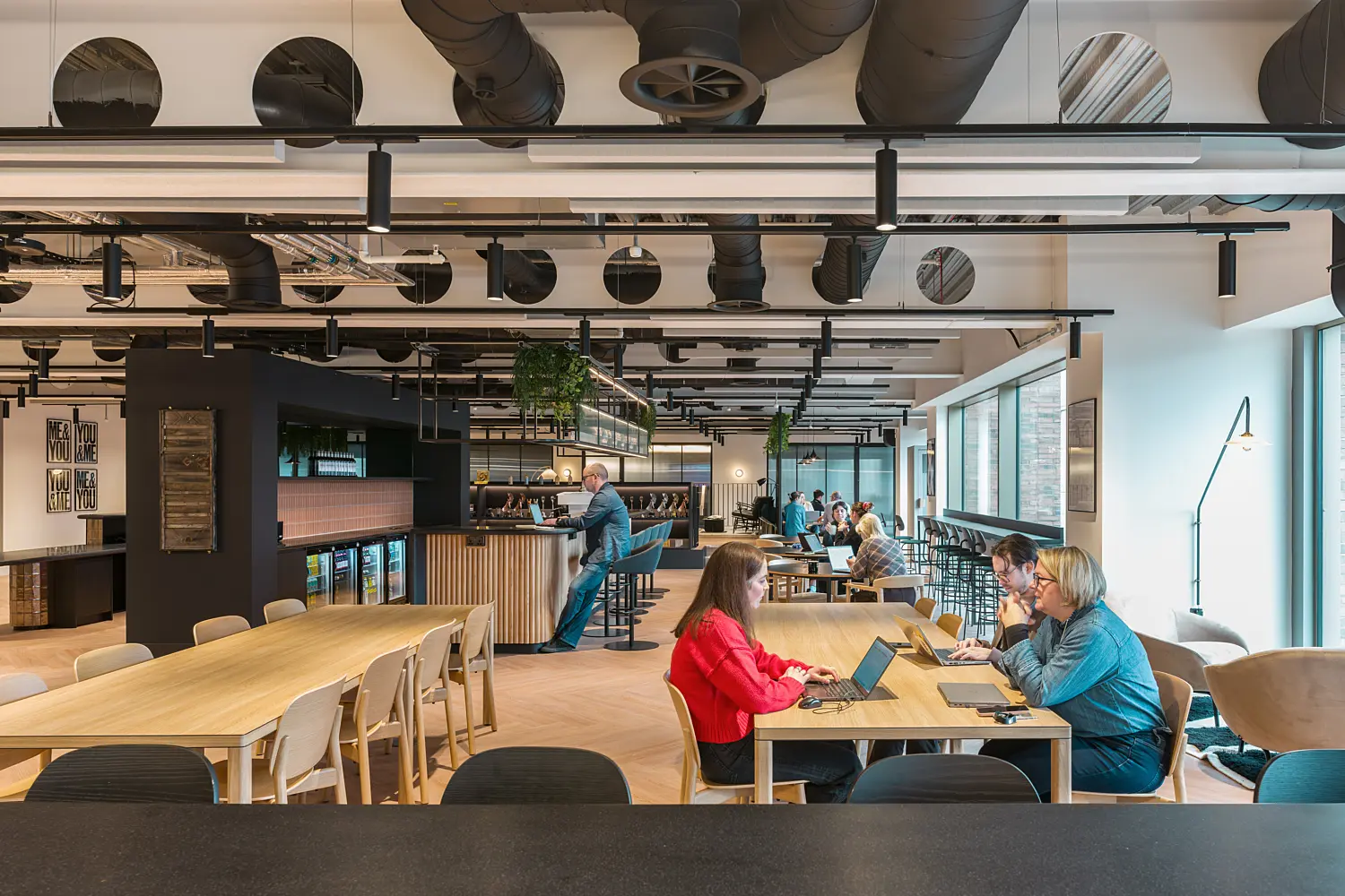 Collaborative workspace within a modern office, featuring long communal tables, soft seating and a central café-style bar area. People are working on laptops and meeting at tables throughout the space. The interior includes light wood flooring, exposed ceiling services, black suspended lighting, large windows providing natural light and integrated planting above the bar. The open-plan layout blends informal meeting, dining and work areas.