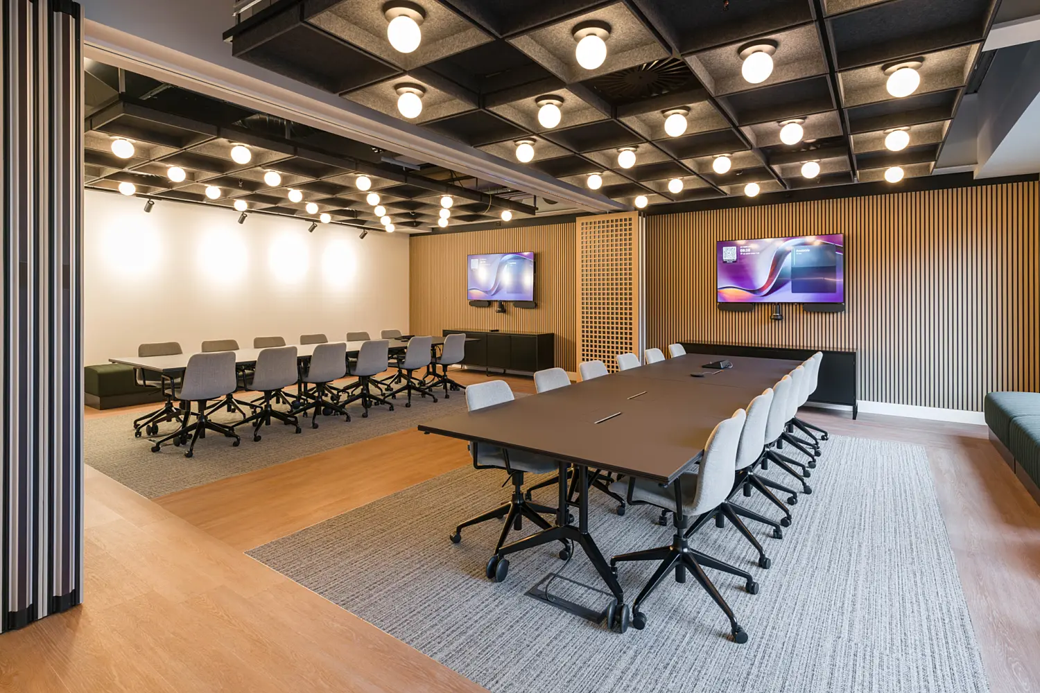 Large multi-purpose meeting and training room with two groups of conference tables surrounded by task chairs on carpeted areas. The space features a black acoustic ceiling with a grid of exposed light fittings, timber slatted wall panelling and two wall-mounted presentation screens. Light wood flooring, flexible furniture arrangements and integrated audiovisual technology create a setting suitable for meetings, workshops and collaborative sessions.