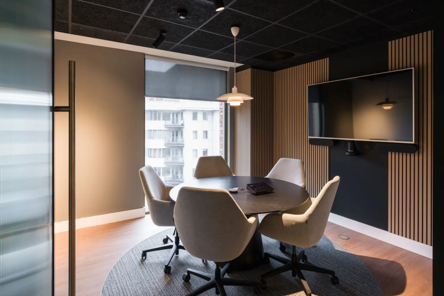 Small meeting room with a round table surrounded by four upholstered task chairs positioned on a circular rug. A large wall-mounted display screen and video conferencing equipment are installed on a dark feature wall with vertical timber slats. Natural light enters through a floor-to-ceiling window with a roller blind, while pendant and ceiling-mounted lighting provide additional illumination. The space combines acoustic finishes, warm wood flooring and contemporary furnishings for private meetings and collaboration.