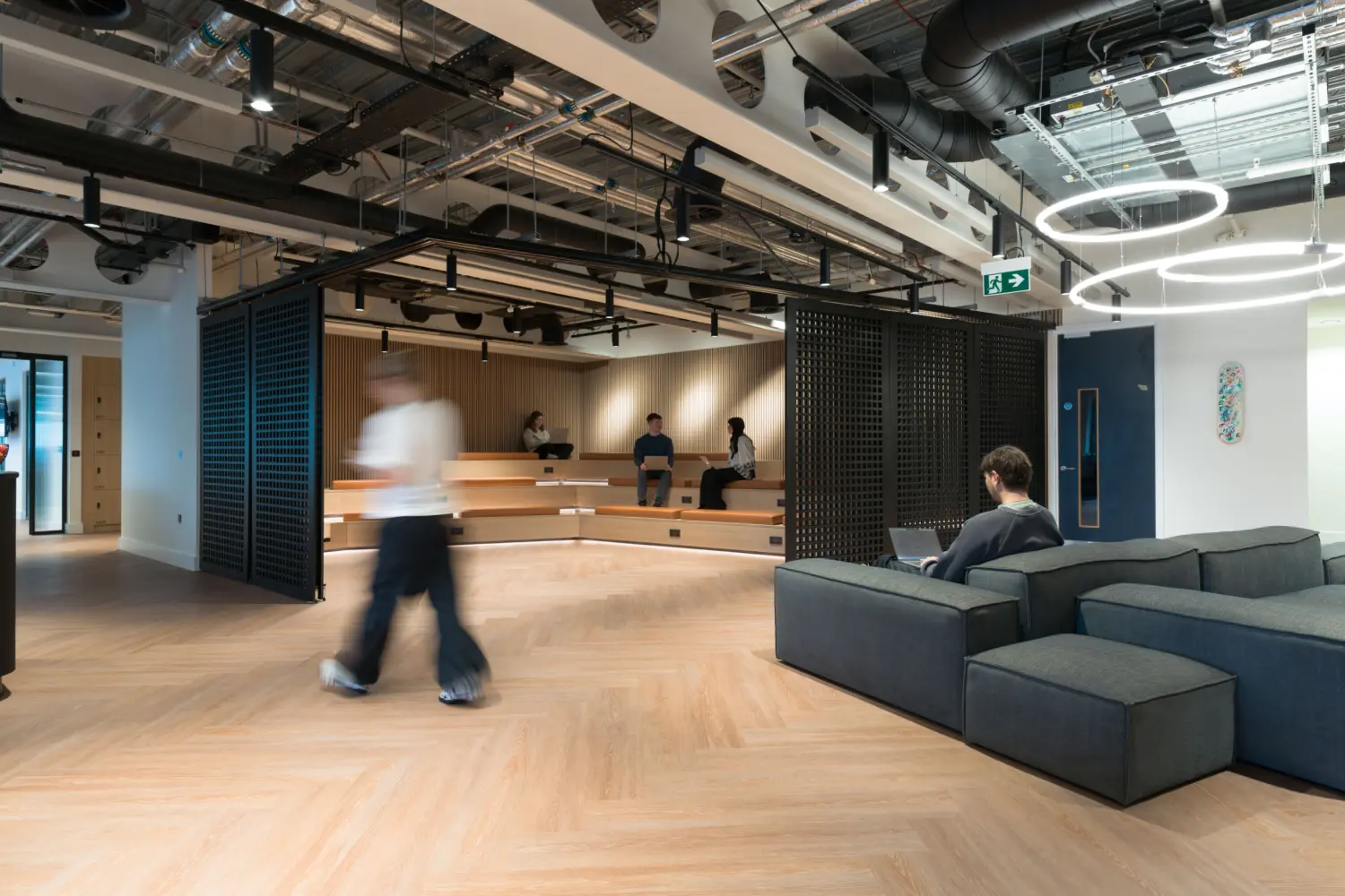 Contemporary office breakout area with light wood flooring, exposed ceiling services and large circular LED pendant lights. Tiered timber seating forms an informal presentation and collaboration space at the centre, where several people are seated. Modular grey lounge seating occupies the foreground, while black lattice screens partially enclose the stepped seating area. A person walks through the open-plan space, which combines social, work and event functions within a bright, modern interior.