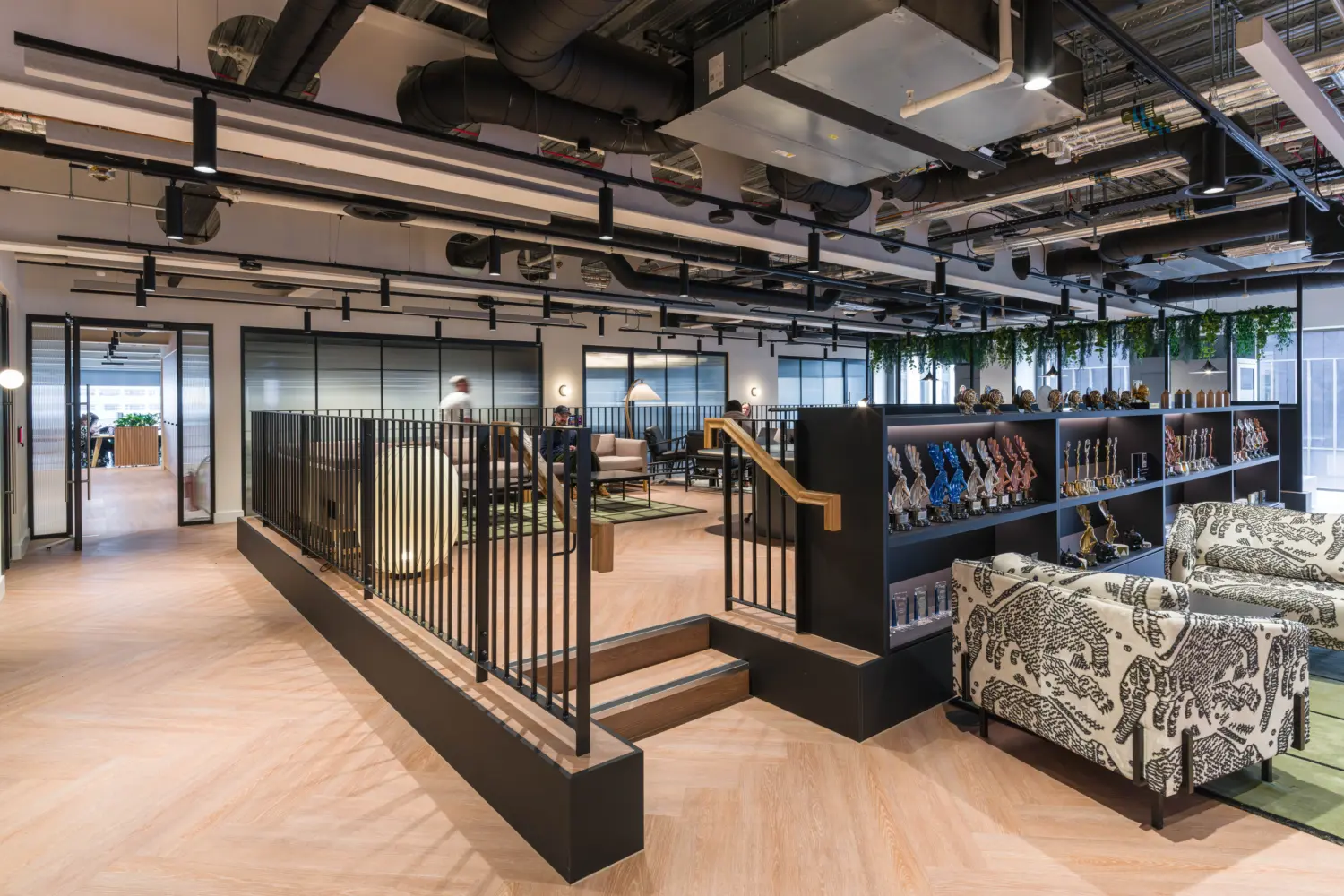 Modern office breakout and lounge area with light wood flooring, exposed ceiling services and suspended black lighting. A raised seating zone with soft furnishings is accessed by a short staircase and enclosed by black metal railings. Open shelving displays a collection of trophies and awards, while patterned armchairs and lounge seating create informal meeting spaces. Glazed meeting rooms line the perimeter, and hanging plants add greenery above the central collaboration area.