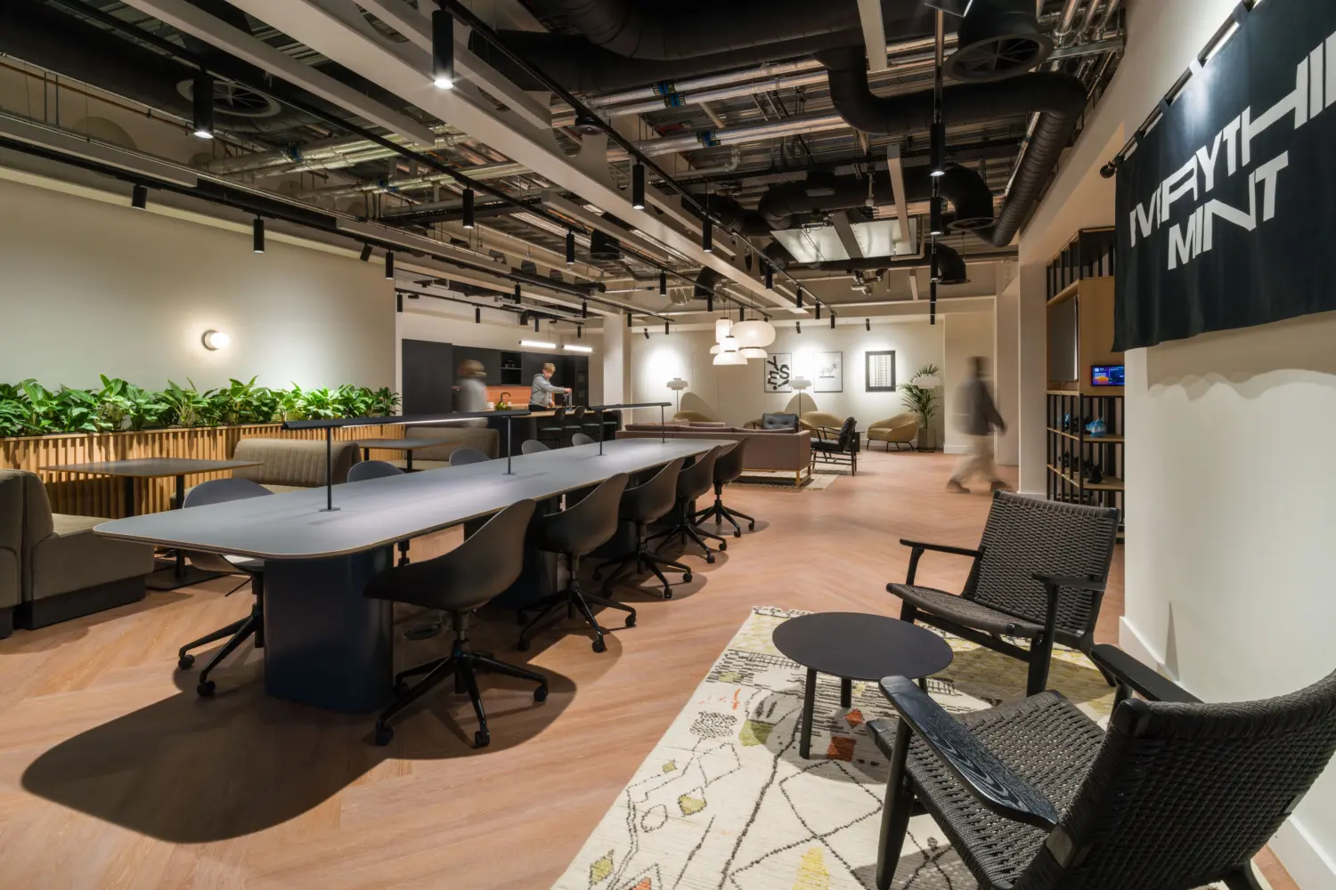 Modern office collaboration space featuring a long shared worktable with task chairs, informal lounge seating and integrated planting. The interior includes light wood flooring, exposed ceiling services and black suspended lighting throughout. A café-style area and breakout seating are visible in the background, while a wall-mounted banner hangs on the right-hand side. The open-plan environment supports both individual work and informal meetings.