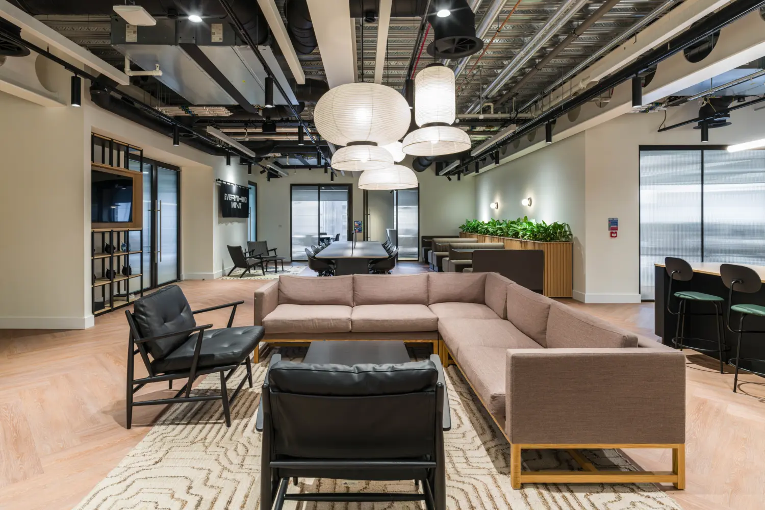 Modern office lounge and collaboration space with a large corner sofa, black lounge chairs and a central coffee table arranged on a patterned rug. Oversized pendant lights hang above a long shared meeting table, while booth seating with integrated planting runs along one side of the room. The interior features light wood flooring, exposed ceiling services and black suspended lighting. Glazed meeting rooms and a bar-style counter are positioned around the perimeter, creating a flexible environment for informal meetings, focused work and social interaction.