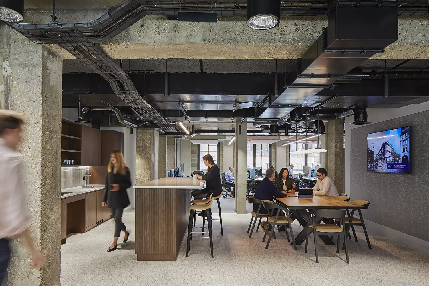 Contemporary office space with an industrial aesthetic, featuring exposed concrete beams and ceiling pipes. The area is illuminated by ample natural light from large windows. The layout includes a communal workspace with tables and chairs, a kitchen area with countertops and cabinetry on the left, and a wall-mounted television displaying an architectural image on the right. The design blends functionality with an industrial style, encouraging collaboration and informal interaction.