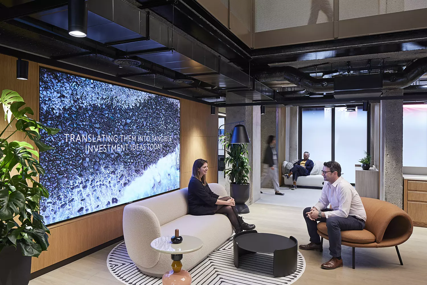 Modern office lounge area featuring a curved beige sofa and a brown chair arranged for informal conversation. A large digital screen on the left wall displays an abstract image with the text 'TRANSFORMING THE WAY THE WORLD WORKS.' The space includes an open ceiling with exposed pipes and lighting fixtures, decorative plants, and large windows that provide natural light. The design blends contemporary comfort with industrial elements, creating a welcoming and innovative atmosphere.