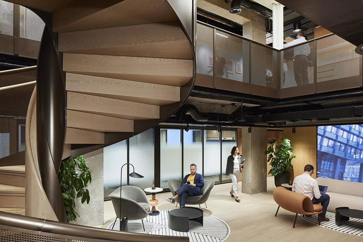 Modern office space featuring a large, central spiral staircase made of wood and metal. The open-plan layout includes high ceilings and expansive windows that let in abundant natural light. The interior design blends industrial and natural materials, with light wood flooring, black chairs around a round table, a small sofa set near the windows, and potted plants adding greenery. The overall aesthetic is contemporary, with clean lines and a minimalist yet inviting atmosphere.