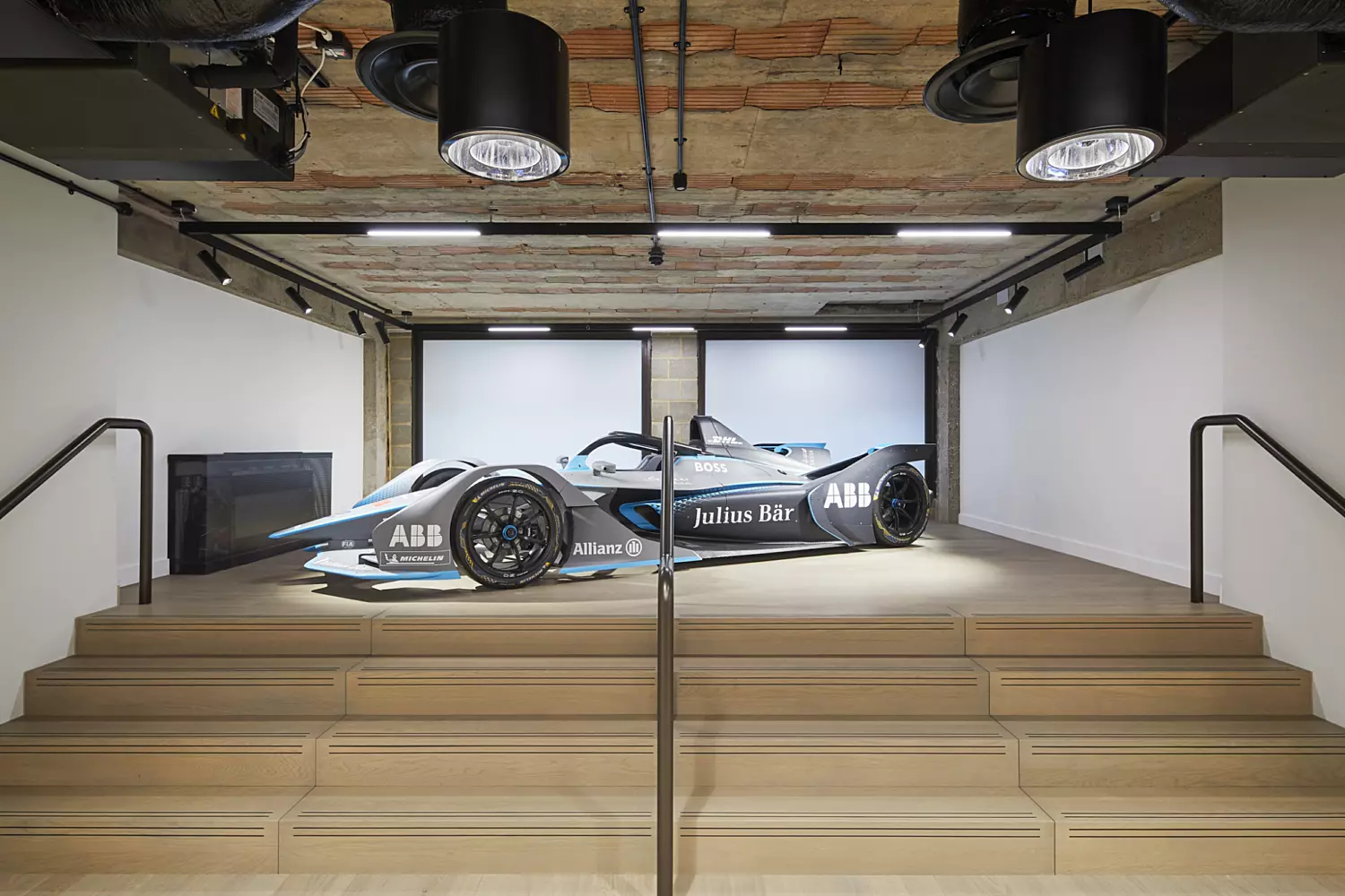 ndoor showroom space featuring a sleek, modern race car on a raised platform. The environment includes industrial-style design elements such as exposed brick ceilings and utilitarian lighting fixtures. The space is clean and minimalistic, emphasizing the car as the central display. The overall aesthetic combines modern exhibition design with raw, industrial architecture.
